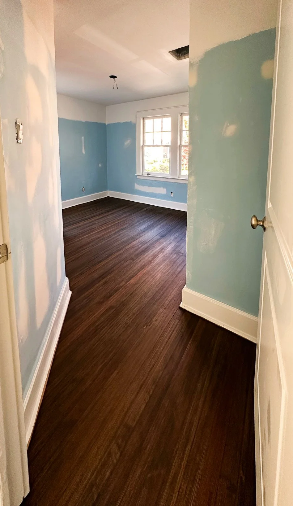 A room under renovation with blue painted walls, a window, dark hardwood floor, and an unfinished ceiling with a hole.