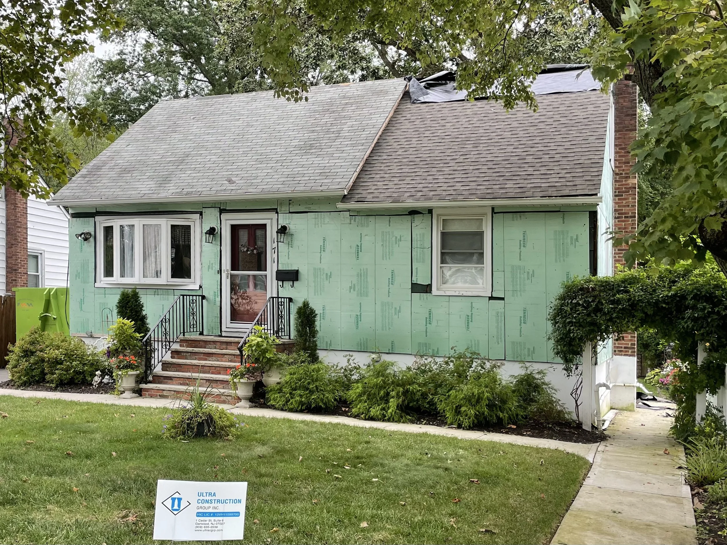A house under renovation with green sheathing siding, a brick front porch with steps, black railings, and a side walkway. The roof is partially covered with a tarp, and there are trees and bushes around the house.