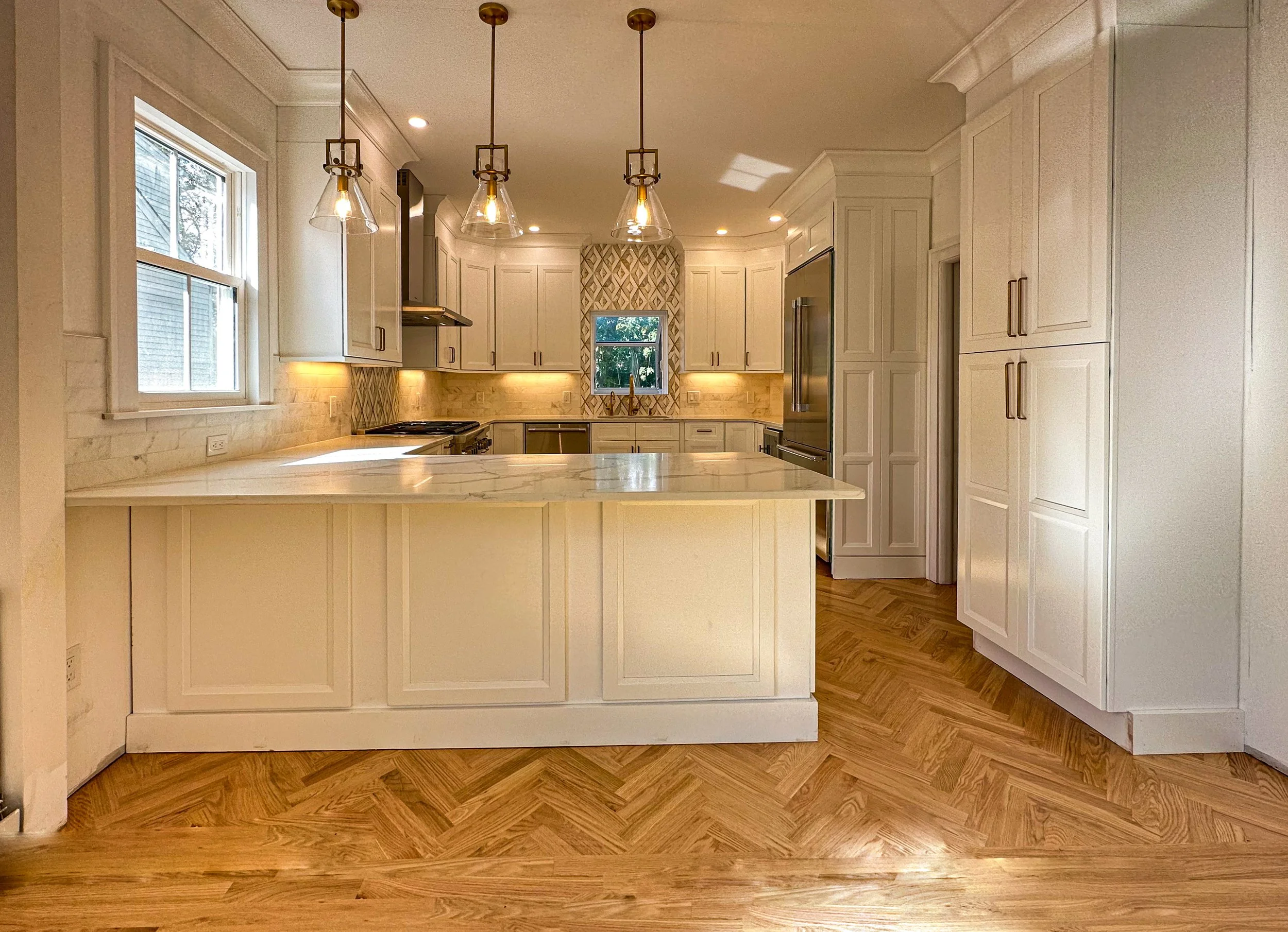 A spacious, modern kitchen with white cabinets, a marble island, hardwood floors, and pendant lighting. There are windows above the sink and a stainless steel refrigerator.