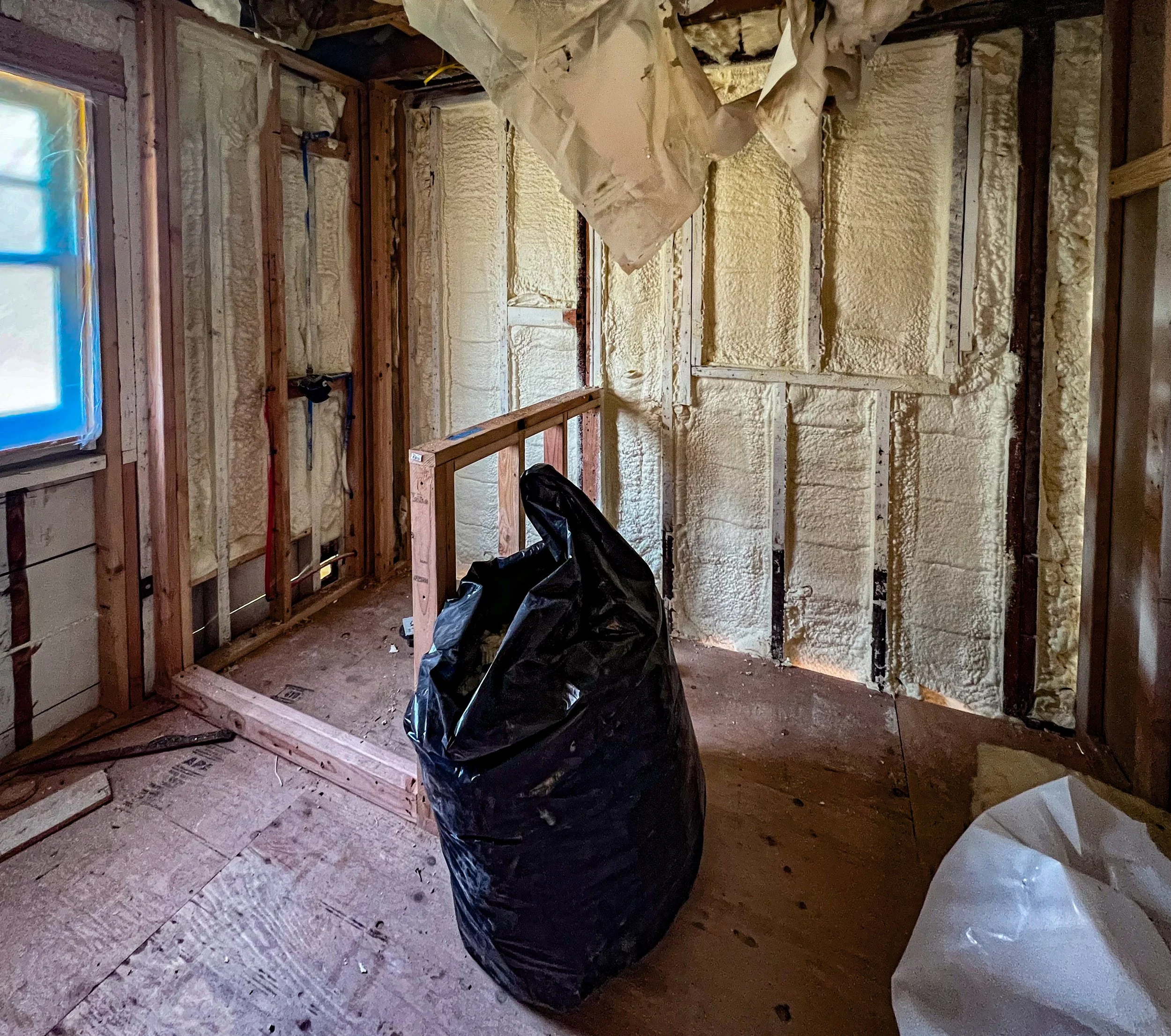 Room under construction with exposed wall studs, spray foam insulation on walls, and a window covered with blue plastic. There is a large black trash bag and a white bag on the unfinished wooden floor.
