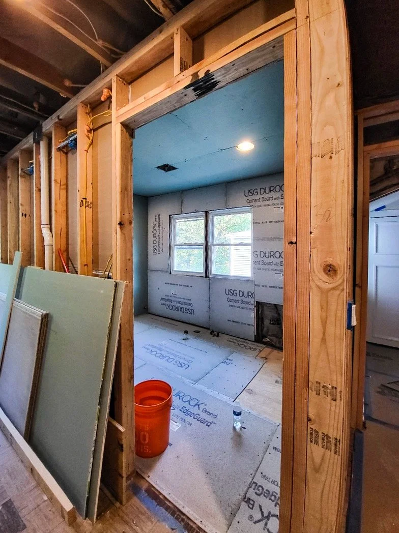 Interior view of a room under construction with framed wooden walls, a window, and construction materials on the floor.