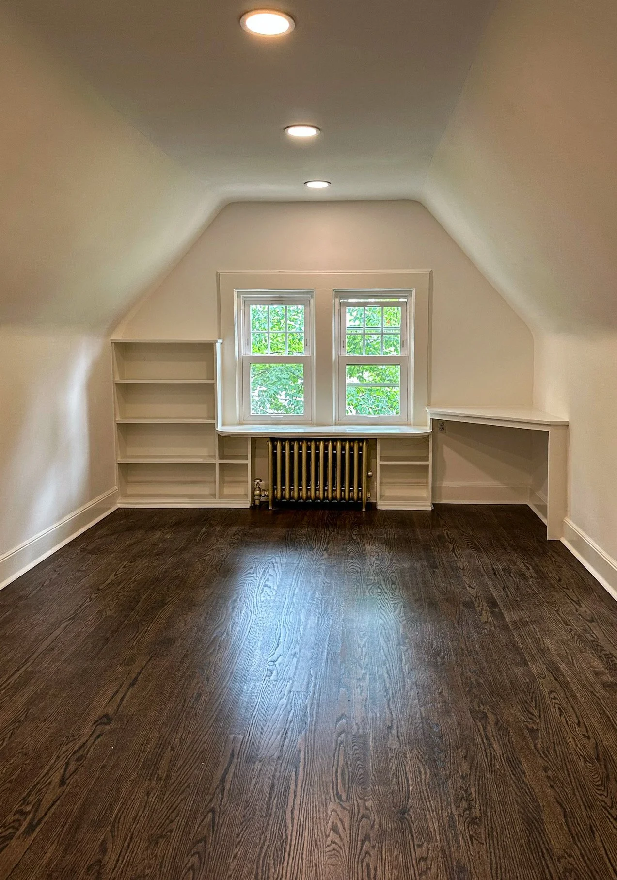 Empty attic room with dark hardwood floors, white built-in shelves, a radiator, and two windows with green trees outside.