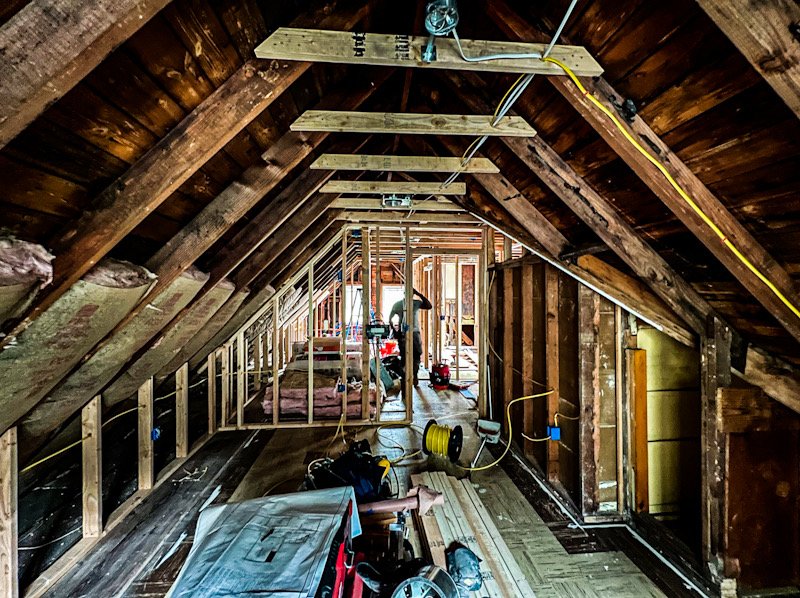 Attic construction in progress with exposed wooden framing, insulation, and construction tools, with a worker in the background.