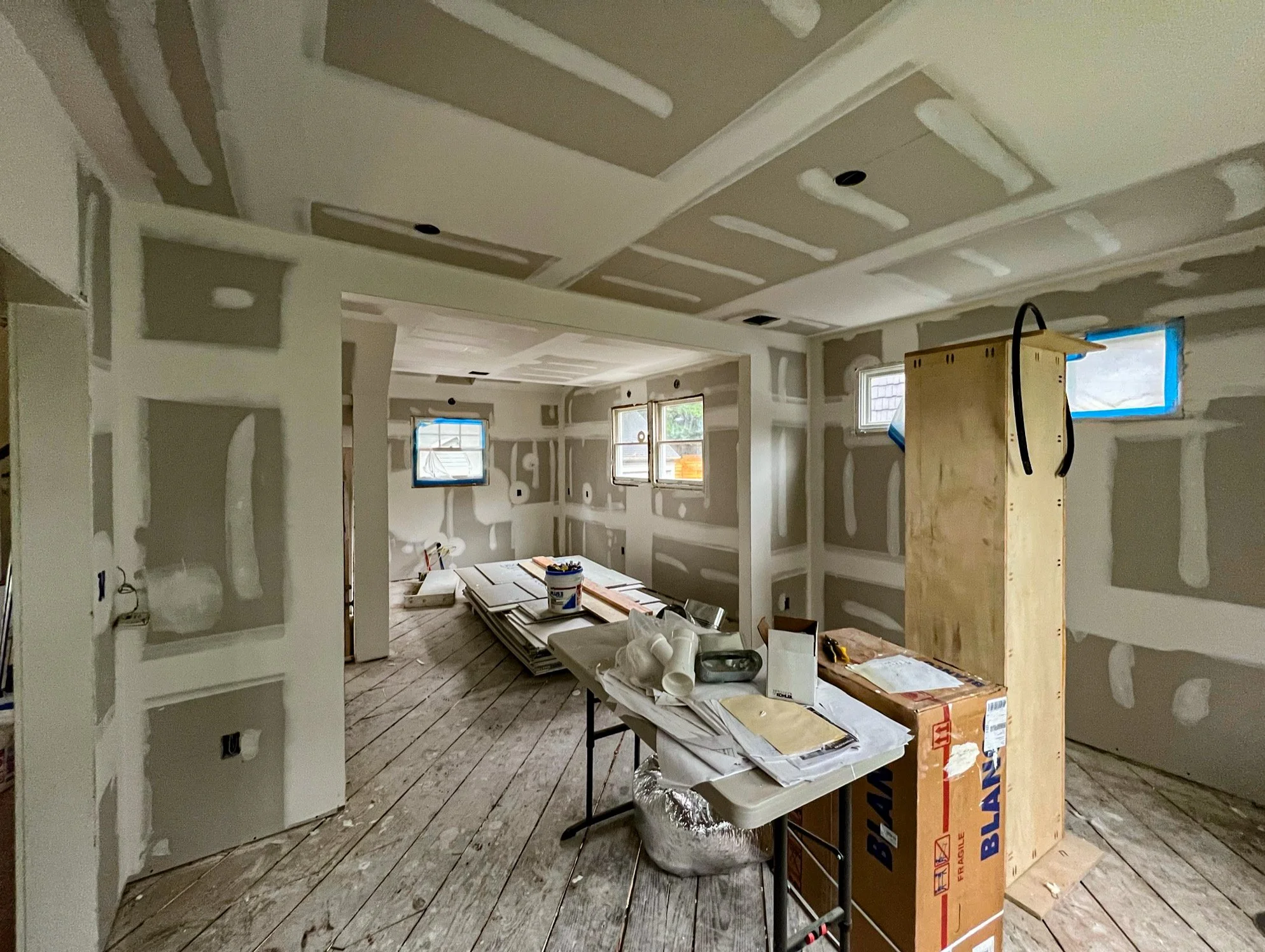 Interior of a room under construction, with drywall installed and taped, construction tools, and building materials scattered on a table and floor, with small windows along the back wall.