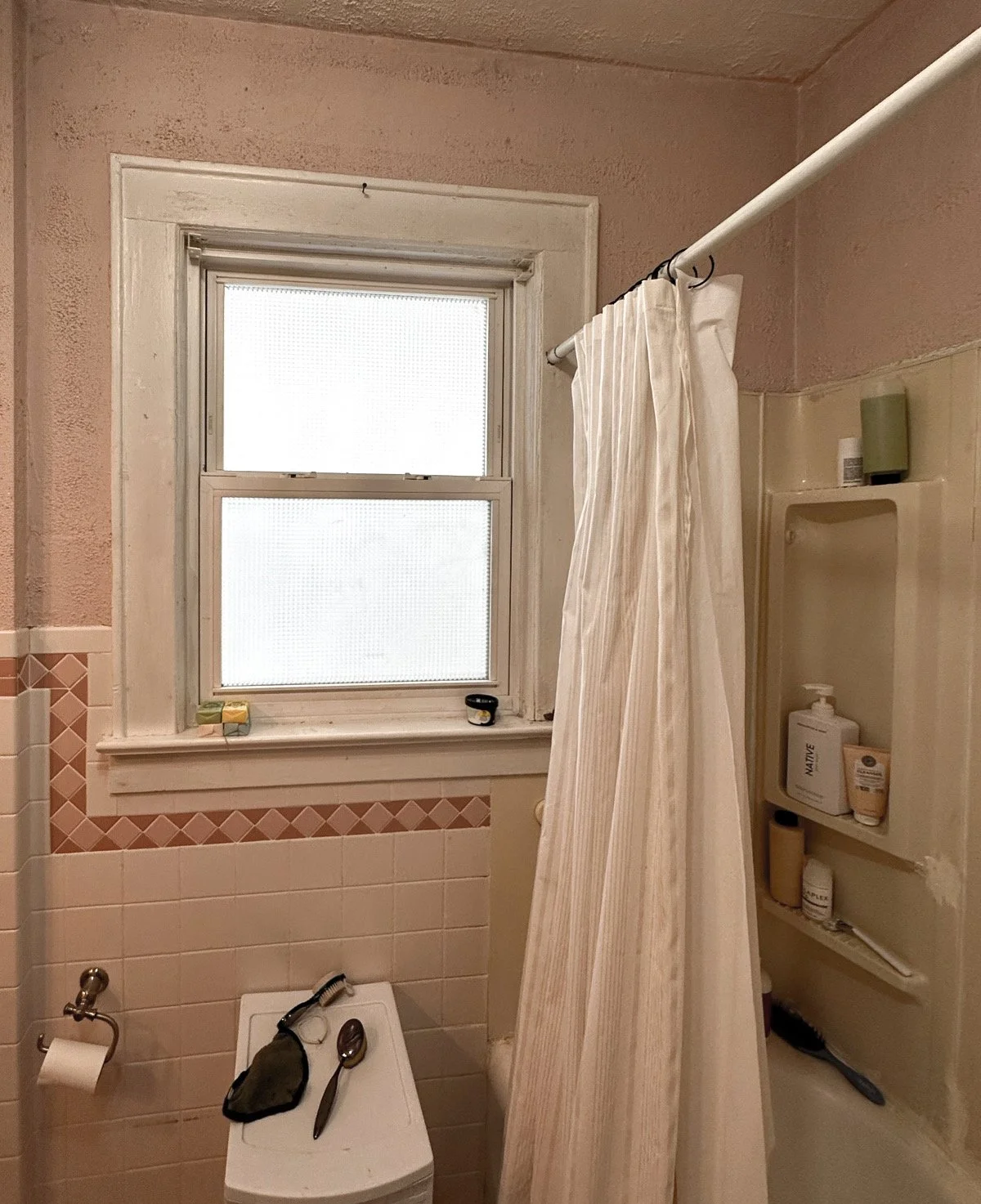 A small bathroom with a window, a shower stall with a curtain, a toilet with a cleaning brush, and a shelf with personal care products.