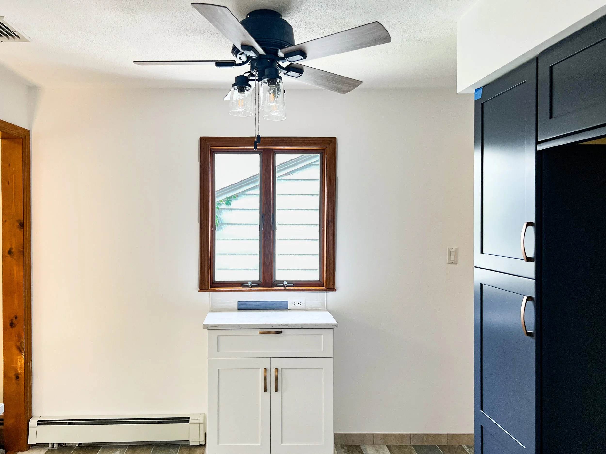 Interior view of a kitchen with a ceiling fan, window, white cabinet, dark blue refrigerator, and a section of wooden trim on a door frame.