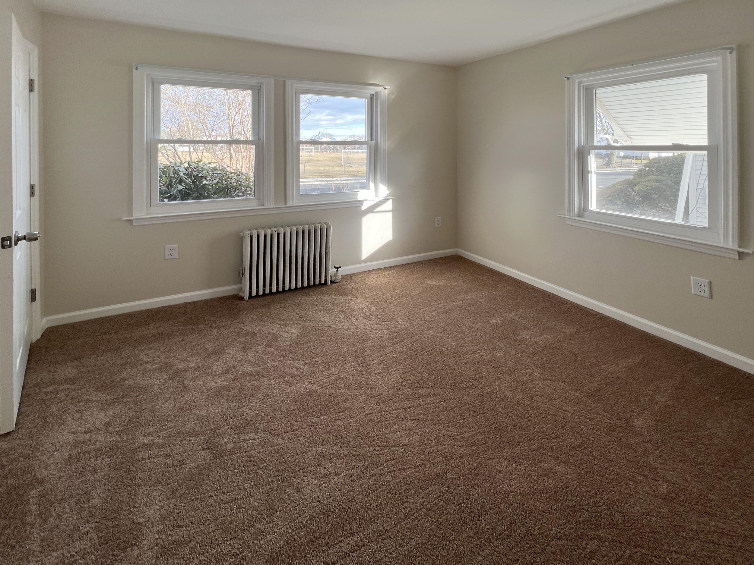 Empty room with beige walls, brown carpet, three windows showing outside view, white trim, and a radiator underneath the window on the left.