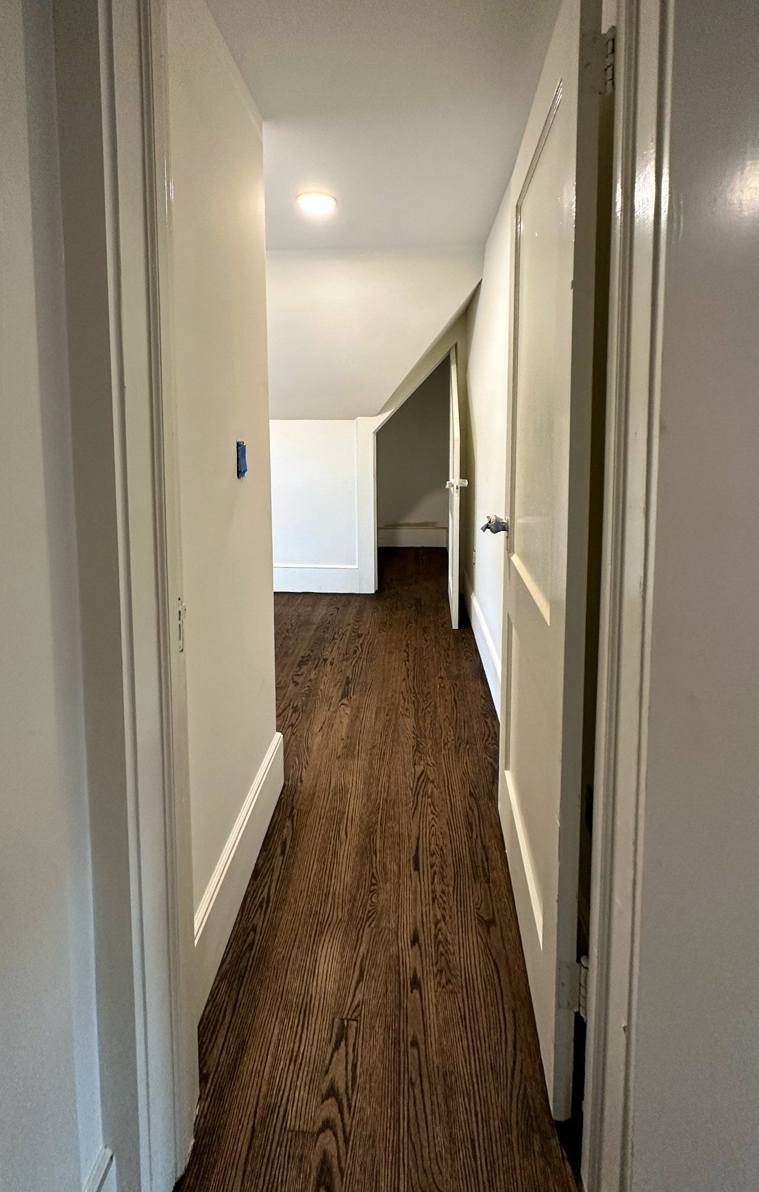 A narrow hallway with wooden floors and white walls, leading to a sloped ceiling with an opening on the right side.