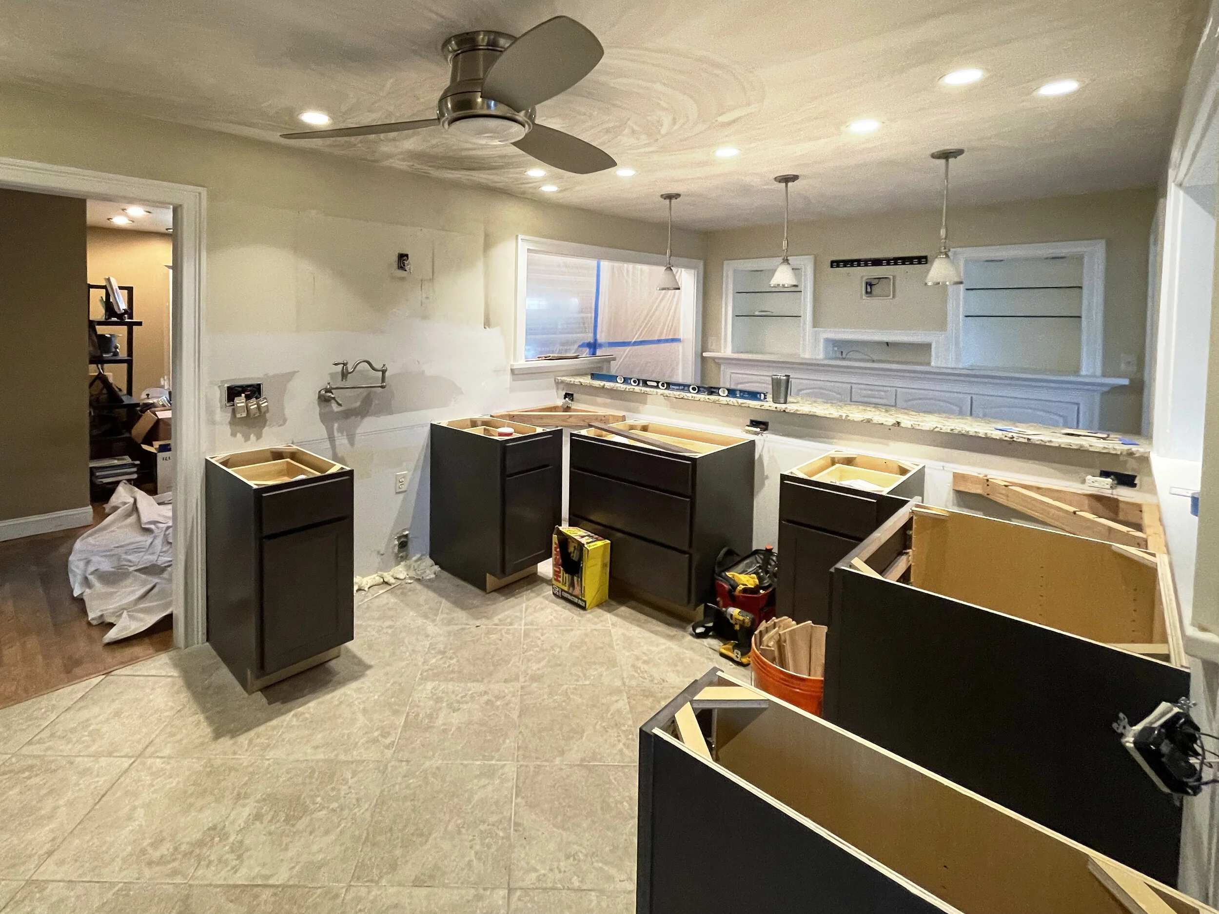 Kitchen undergoing renovation with dark cabinetry, granite countertops, and a ceiling fan, with cabinet boxes and tools visible.
