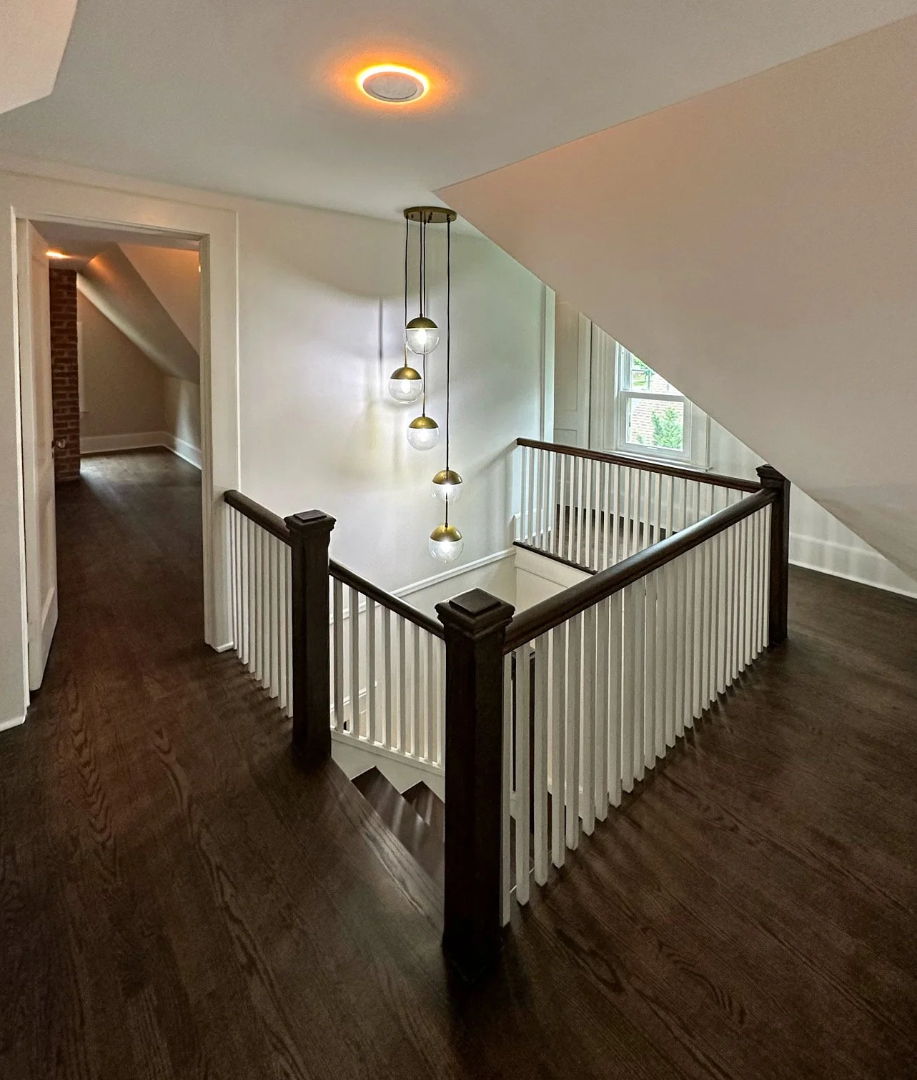 Interior view of a home staircase with dark wooden floors, white balustrades with dark wood handrails, a modern hanging light fixture with multiple glass globes, a window letting in natural light, and an open doorway leading to a room with exposed br