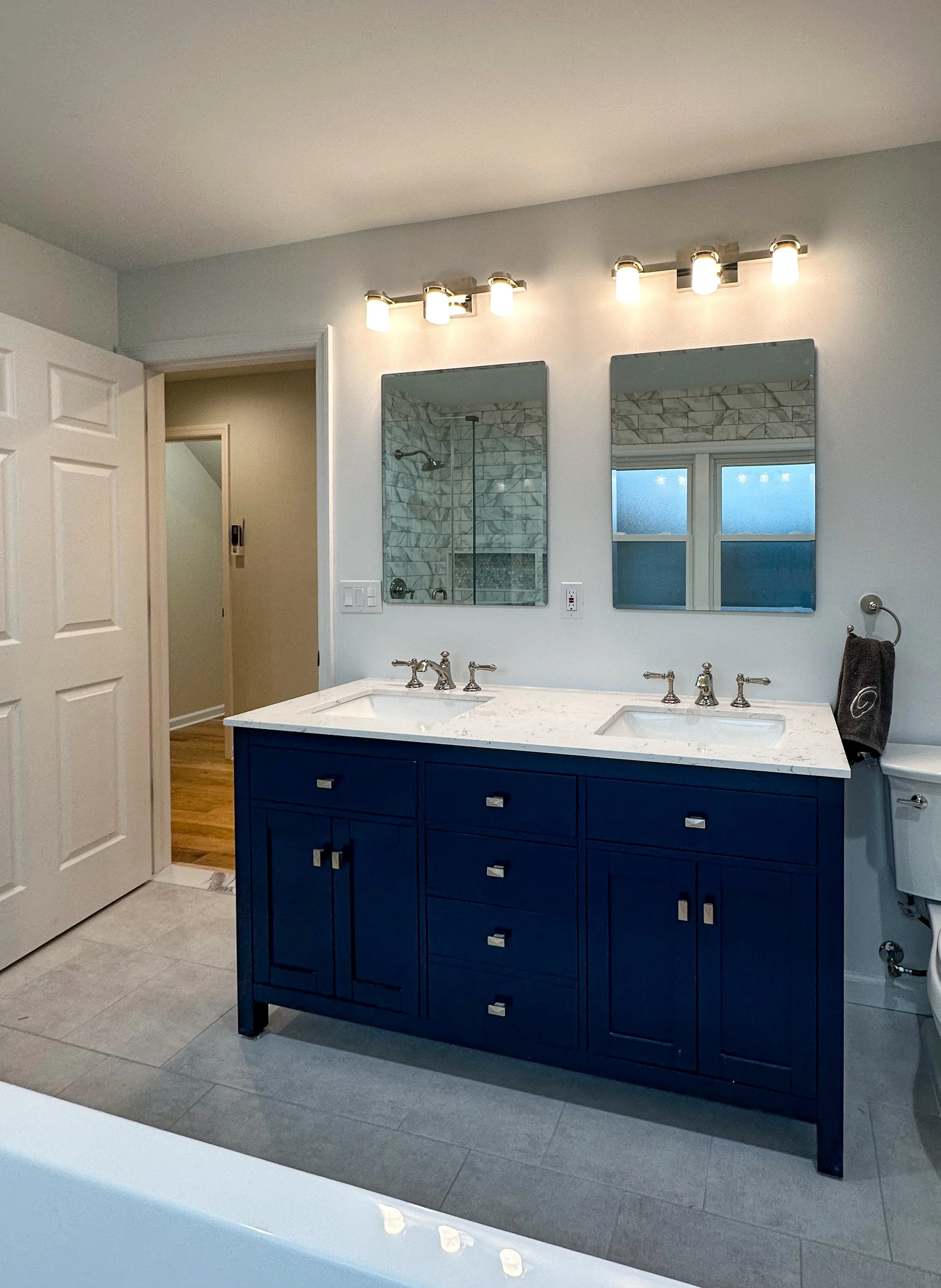 A modern bathroom vanity with a white marble countertop, two undermount sinks, and navy blue cabinets. Two mirrors are mounted above the vanity, with a set of three lights above each mirror. A towel hangs on a ring to the right of the vanity, and a w