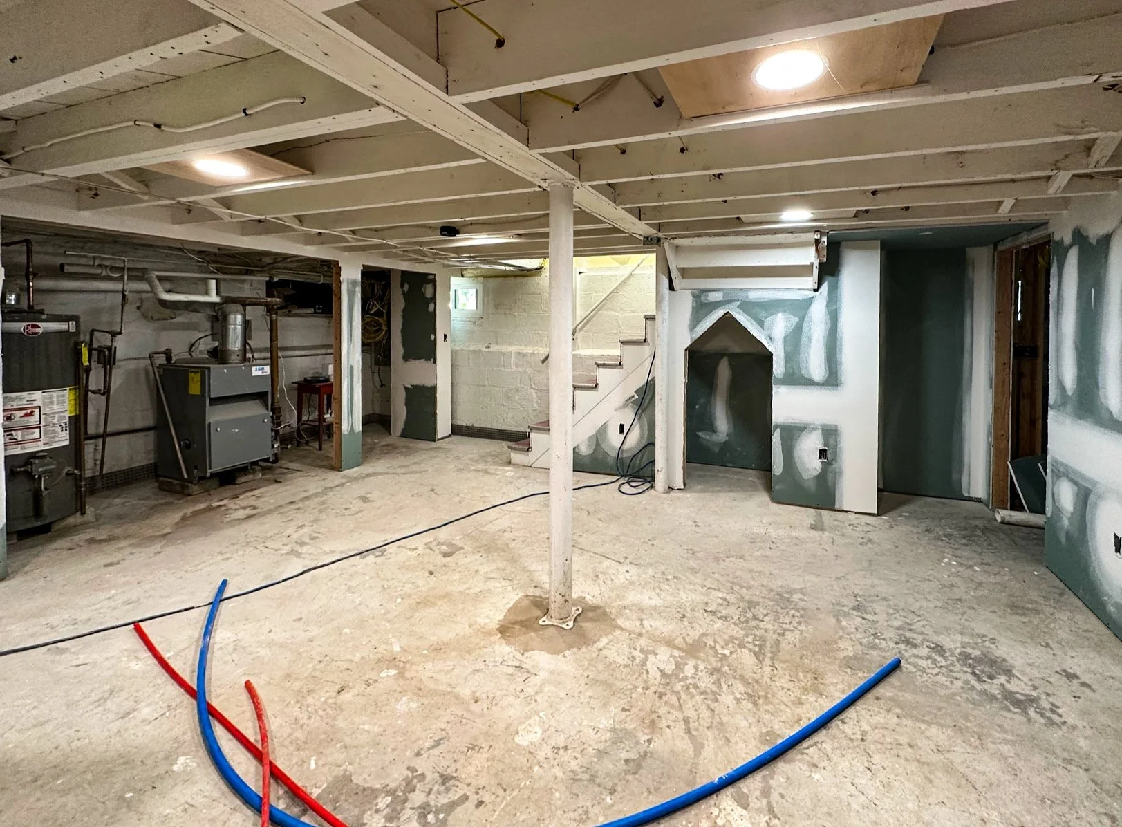 Underground basement with exposed floor and ceiling, electrical and HVAC equipment, drywall installation in progress, and staircase leading to upper level.