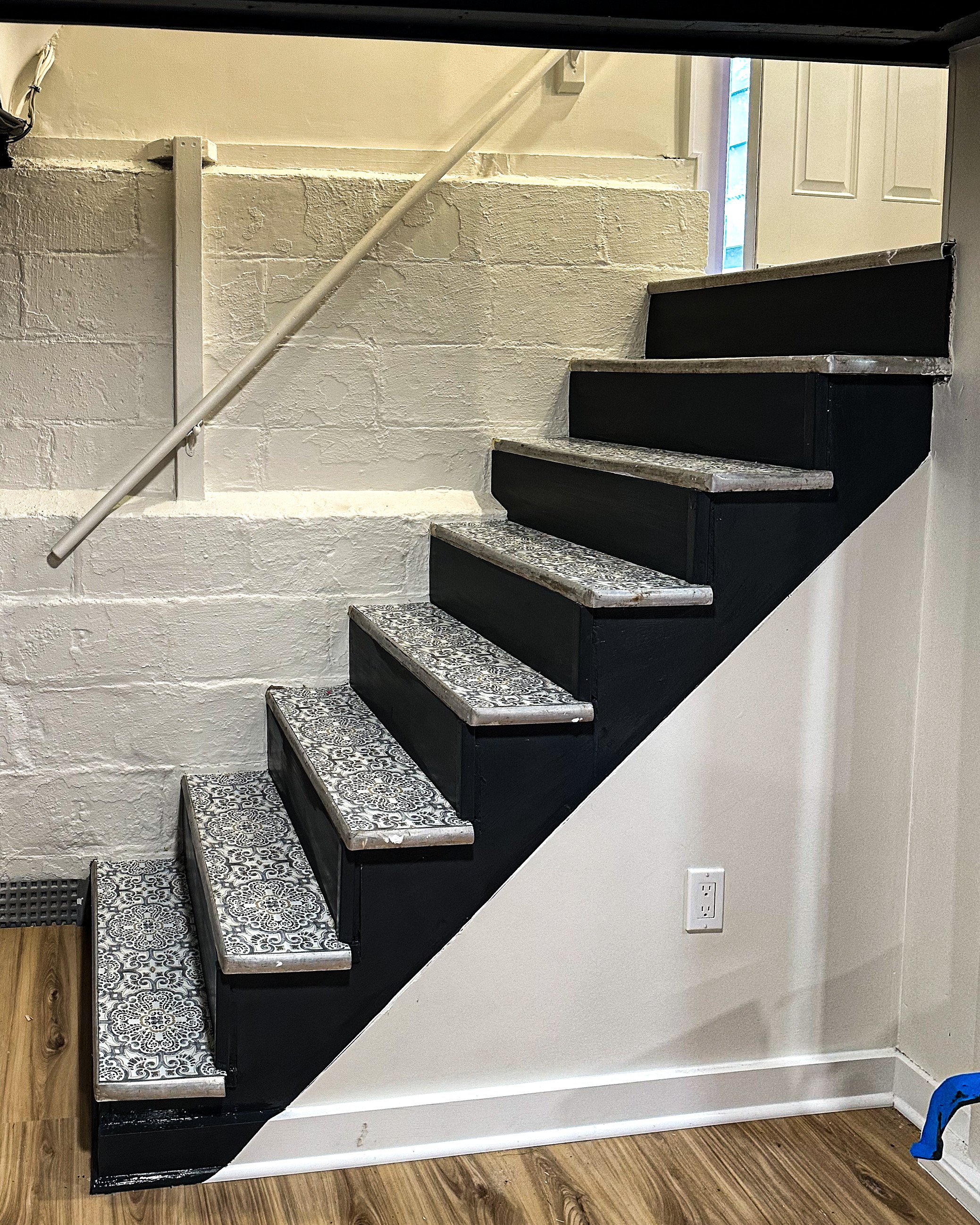 Interior view of a staircase with black painted risers, decorative tiled treads, a white wall, a textured brick wall, and a white railing.