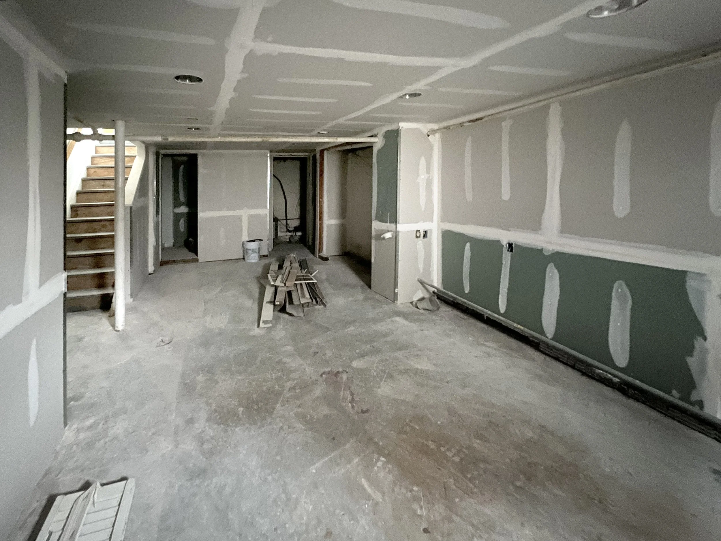 Unfinished basement with drywall installation and exposed concrete floor, stairs visible on left, construction materials in the center, and no furniture or decor.