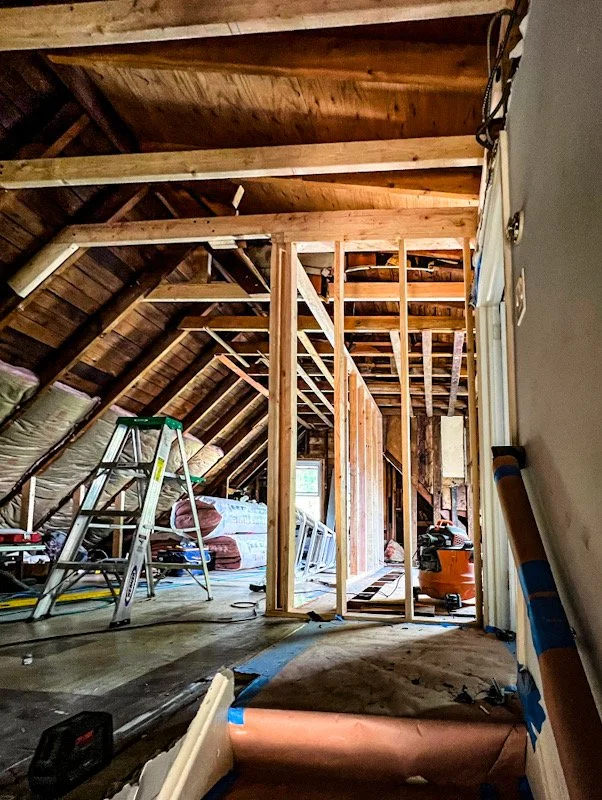 Attic space under renovation with wooden framing, a step ladder, insulation materials, and construction tools visible.