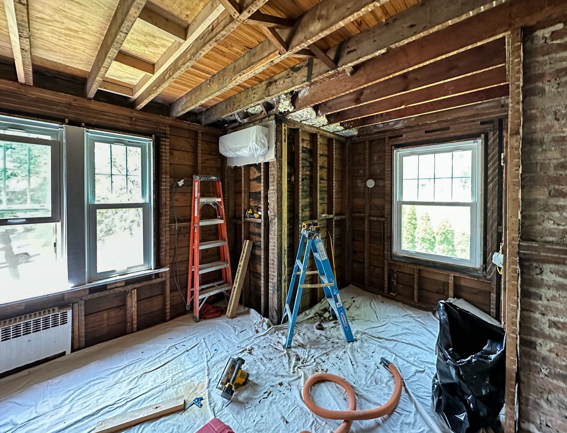 Interior of a room under renovation with exposed wooden framing, construction tools, ladders, plastic covering the floor, and two windows letting in natural light.
