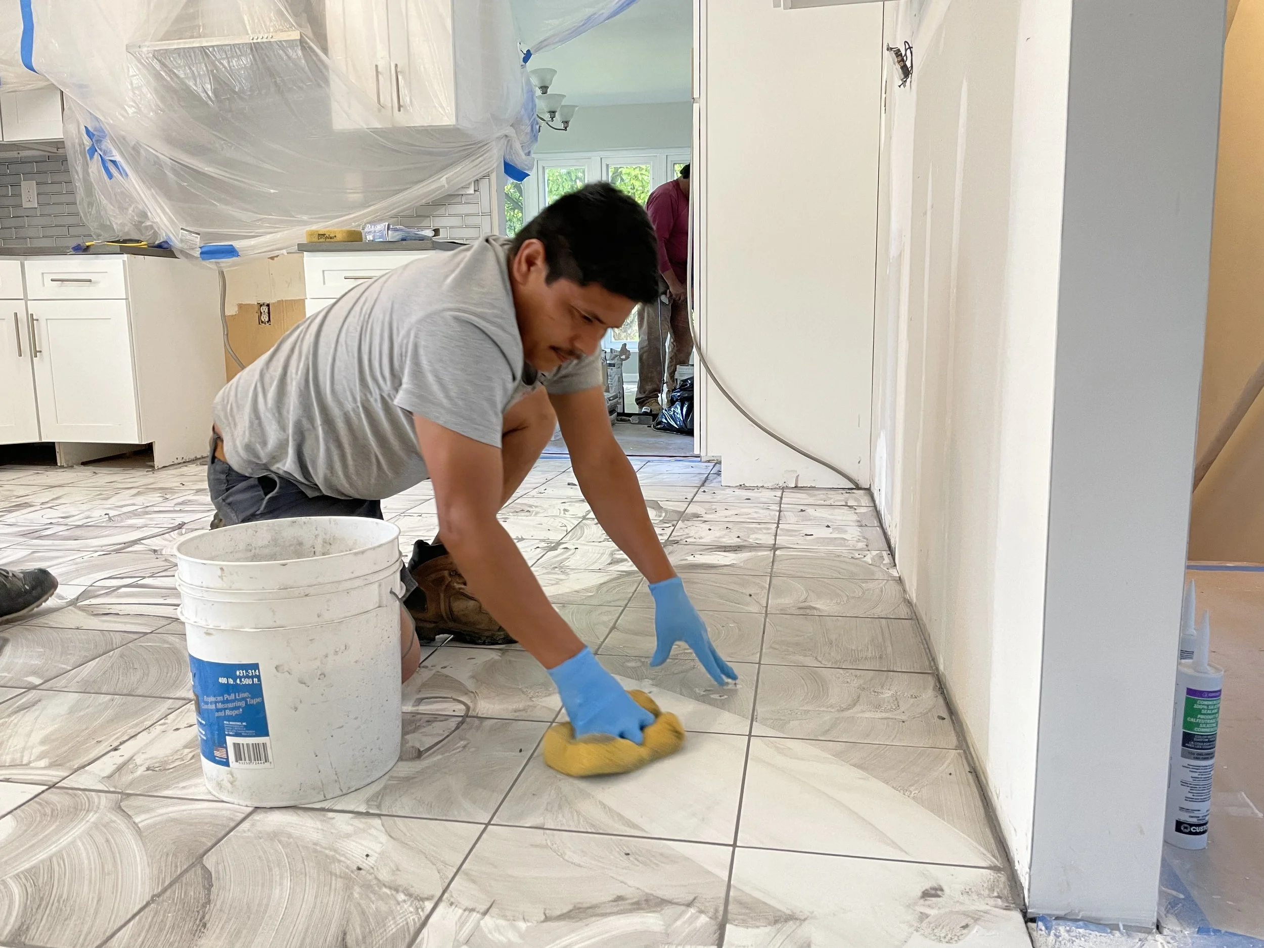 Man installing or cleaning marble tile floor, wearing gloves, kneeling, with construction supplies around, in a kitchen under renovation.