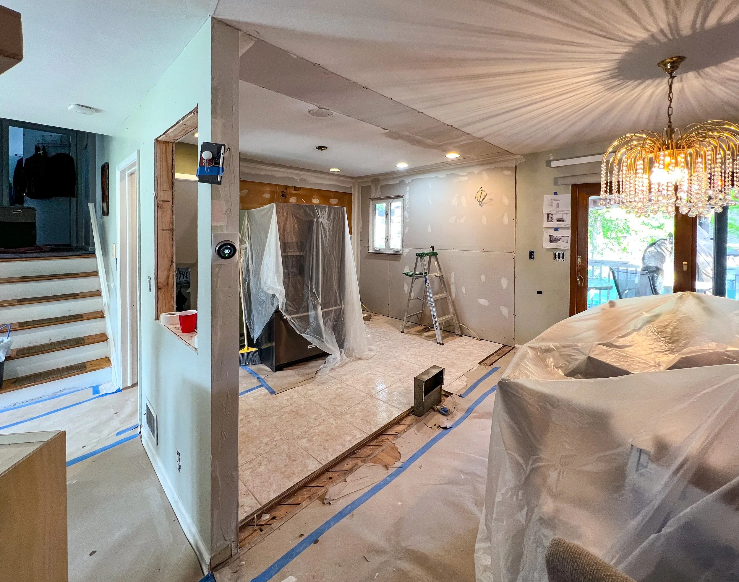 Living room under renovation with drywall work, covered furniture, ladder, and construction tools.