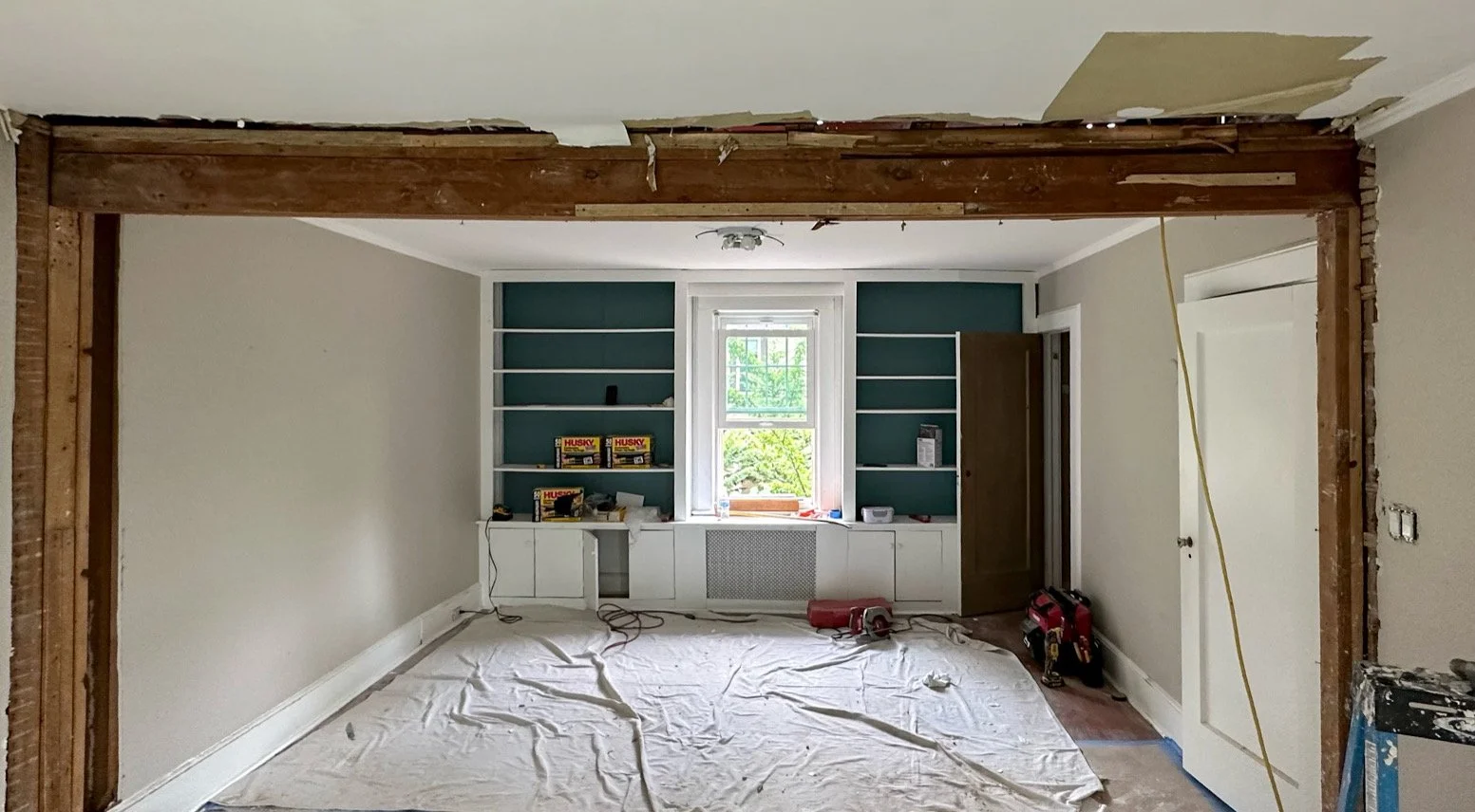 Room undergoing renovation with a large wooden beam framing the doorway, a window with built-in shelves, and construction tools on the floor covered with a drop cloth.