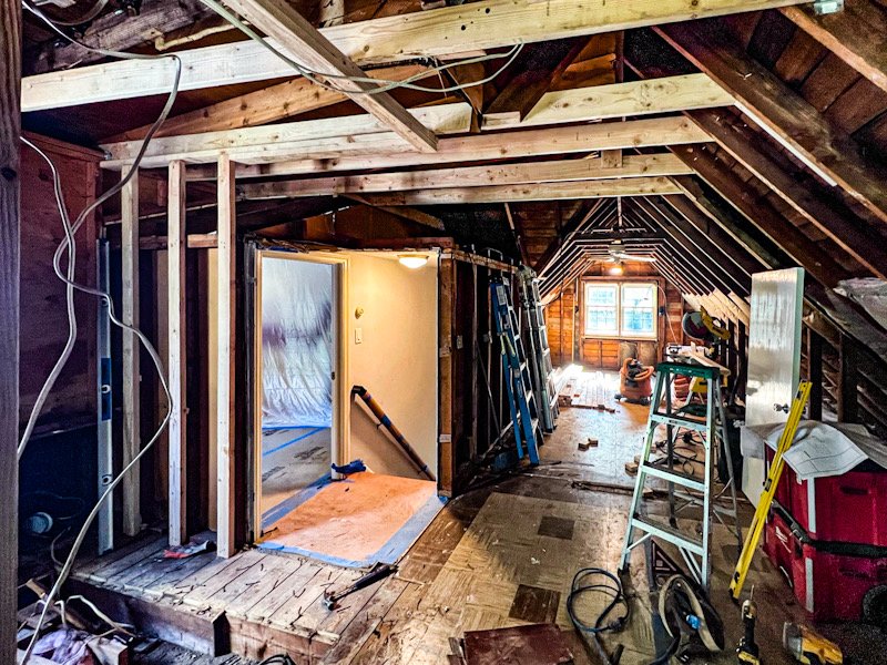 Attic renovation with exposed wooden beams, construction tools, a stepladder, and partially built walls, showing ongoing carpentry work.