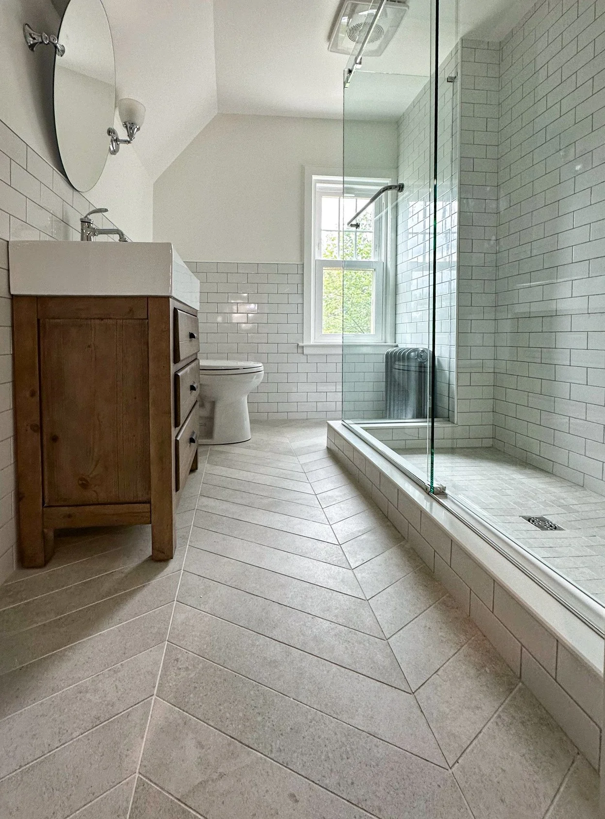 A modern bathroom with a glass-enclosed shower, white subway tile walls, a window with natural light, a wooden vanity with a white sink, a mirror, and a toilet.