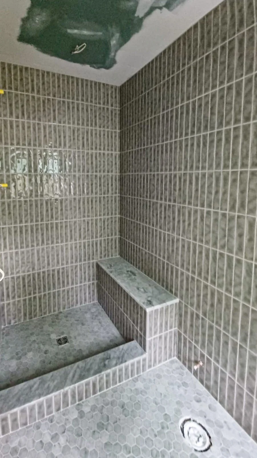 A tiled shower area with small beige tiles on the walls, a built-in bench, a floor drain, and an unfinished ceiling with exposed drywall.