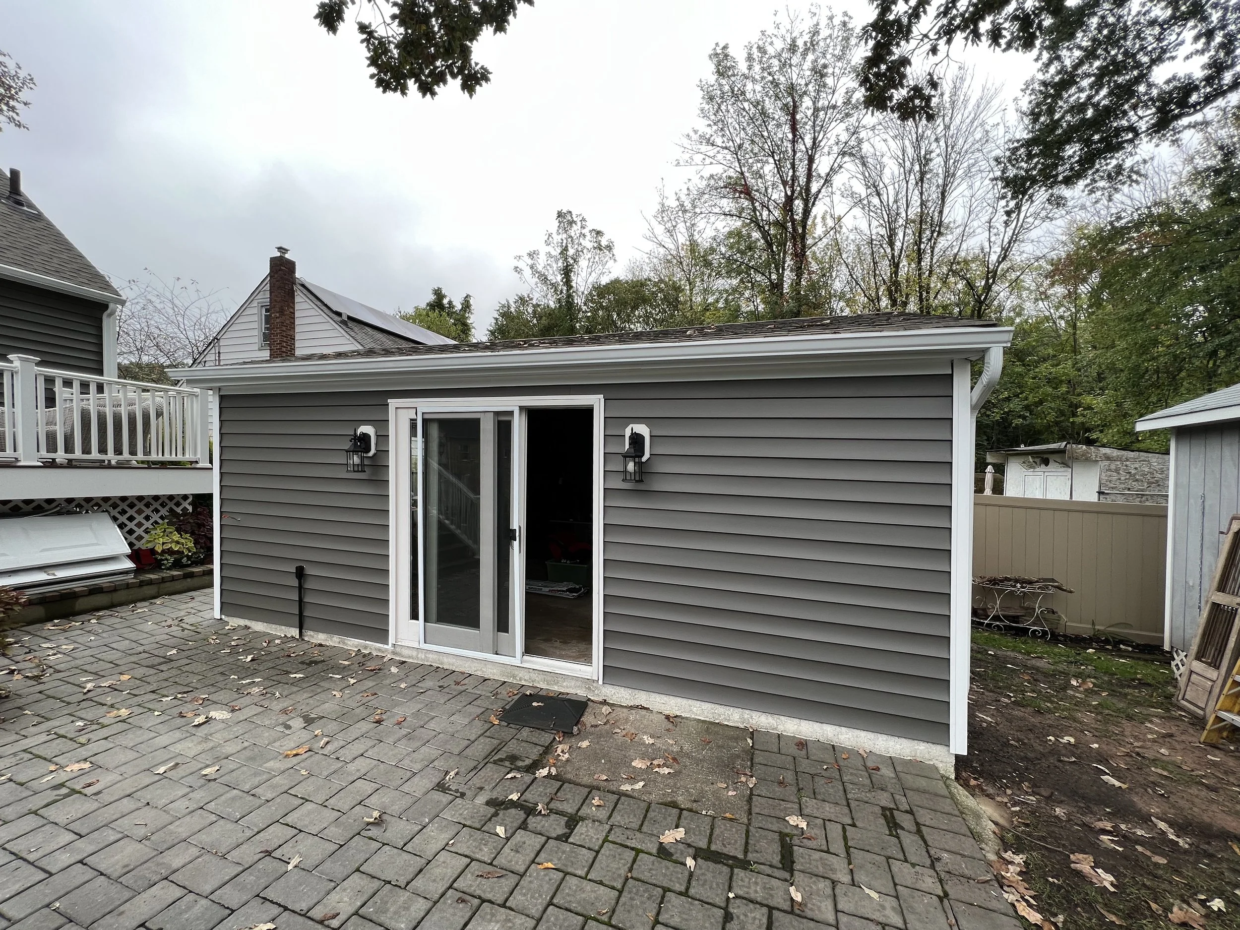 Backyard view of a small gray house or shed with sliding glass door, two outdoor wall-mounted lights, and paved stone patio, with trees and neighboring structures in the background.