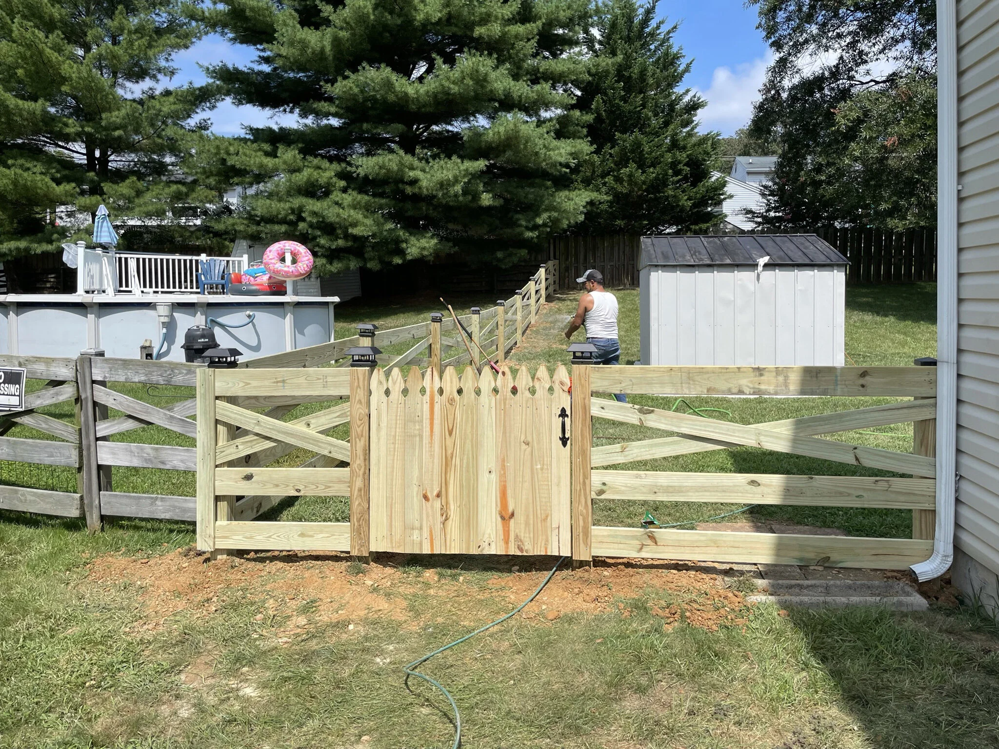A yard with a wooden fence, a man working with garden tools, a shed, a swimming pool with floating toys, and play equipment including a slide and an inflatable ring, surrounded by trees and grass.