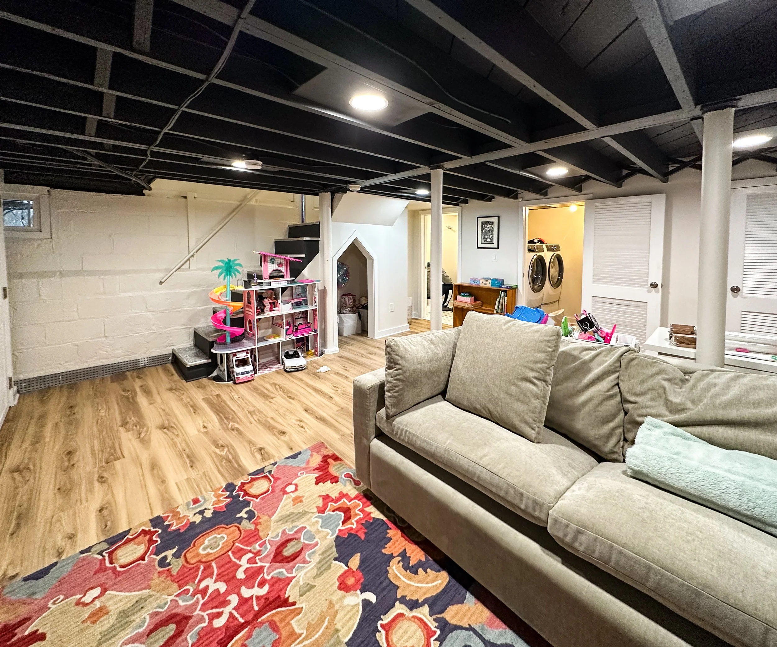 Living room with a beige couch, colorful area rug, toy storage, and laundry area in the background.