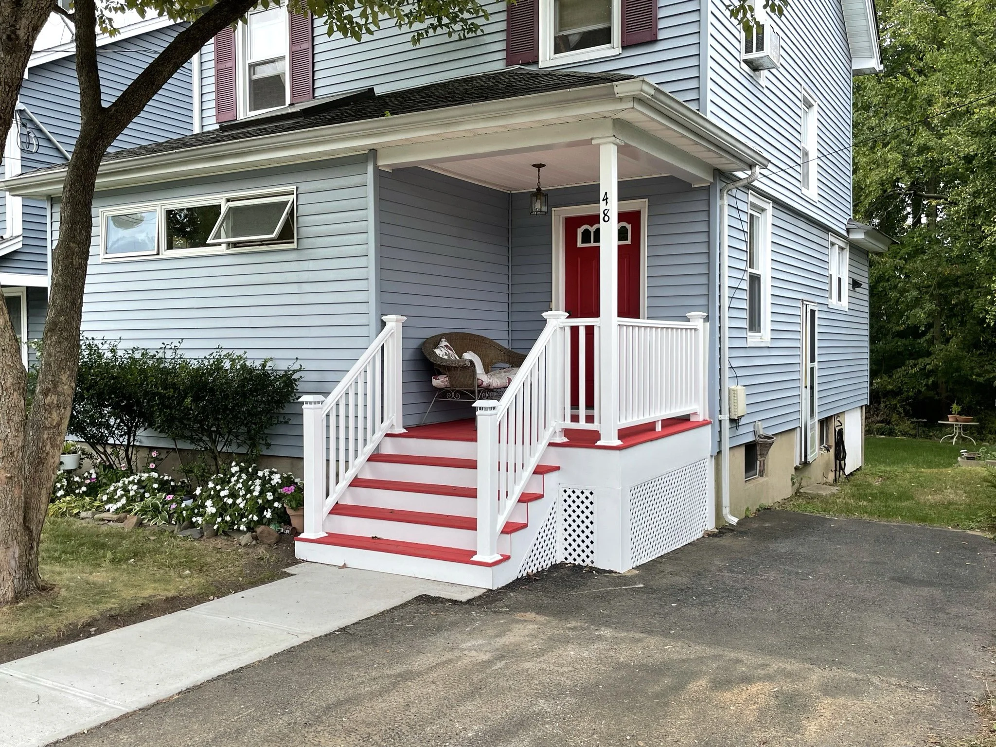 A blue two-story house with white and red accents, a small front porch with white railing, red steps, and a red door. The porch has a wicker loveseat with pillows. There are flower beds with white flowers and green shrubs in front of the house, and a