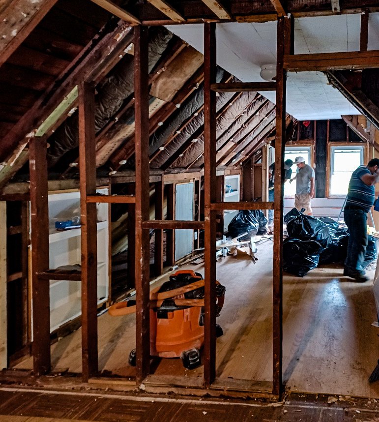 Attic under renovation showing exposed wooden studs, insulation, and workers installing drywall or other materials. There are black trash bags, construction tools, and a vacuum cleaner on the wooden floor.