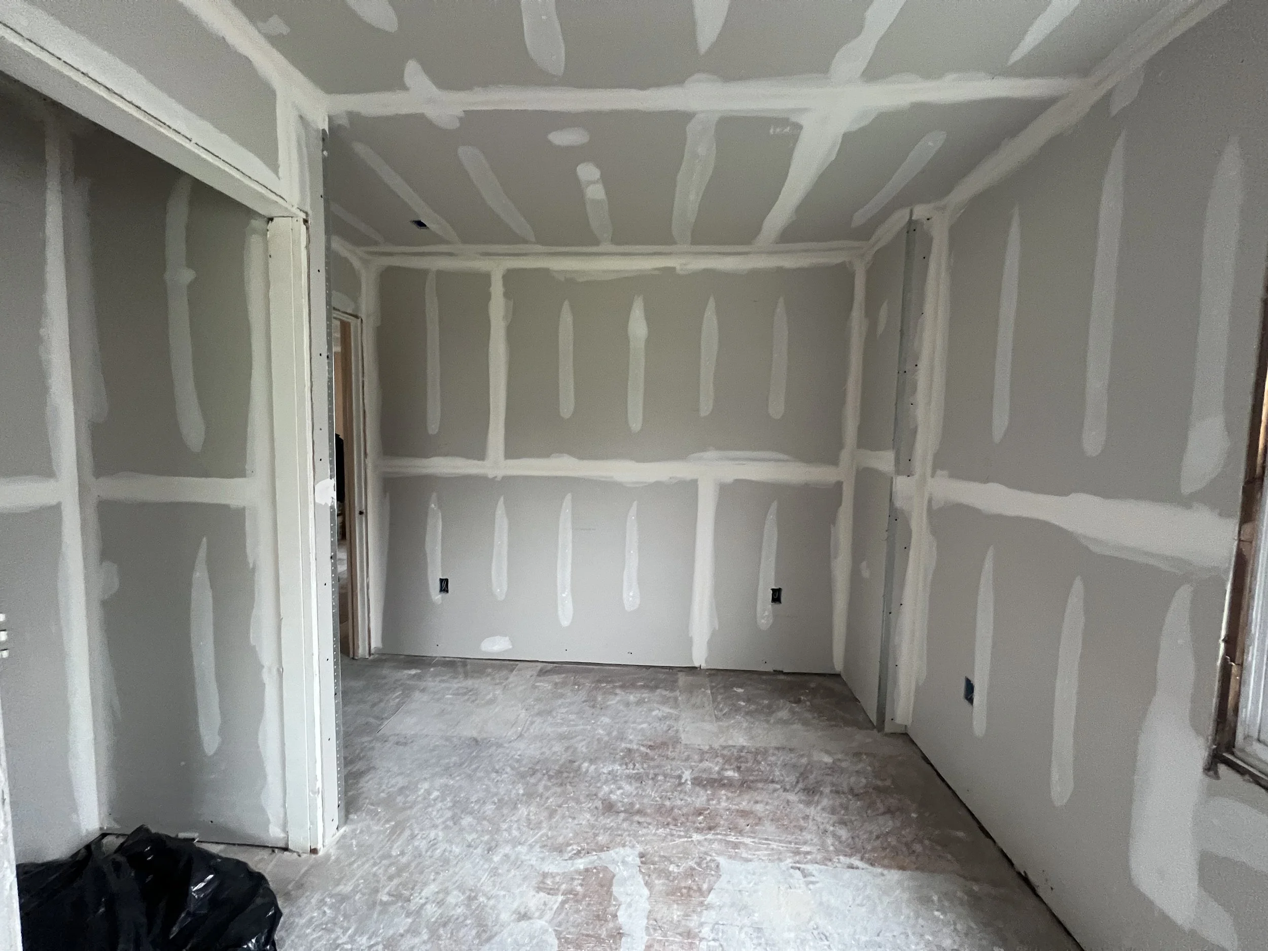 An unfinished room with drywall installed on the walls and ceiling. The drywall has visible joint compound application, and the floor is bare with dust and debris. There is a black object or bag on the floor in the lower left corner, and an open wind
