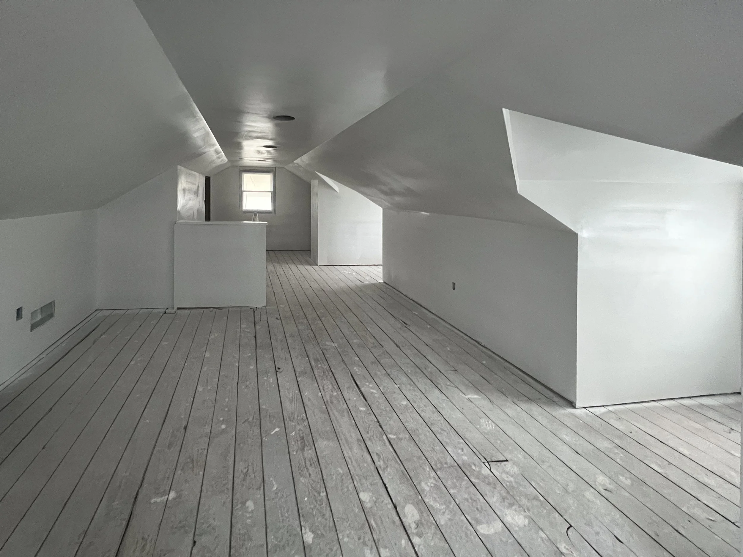 An unfinished attic room with white walls, angled ceilings, and wooden floorboards. There is a small window at the far end allowing natural light to enter.