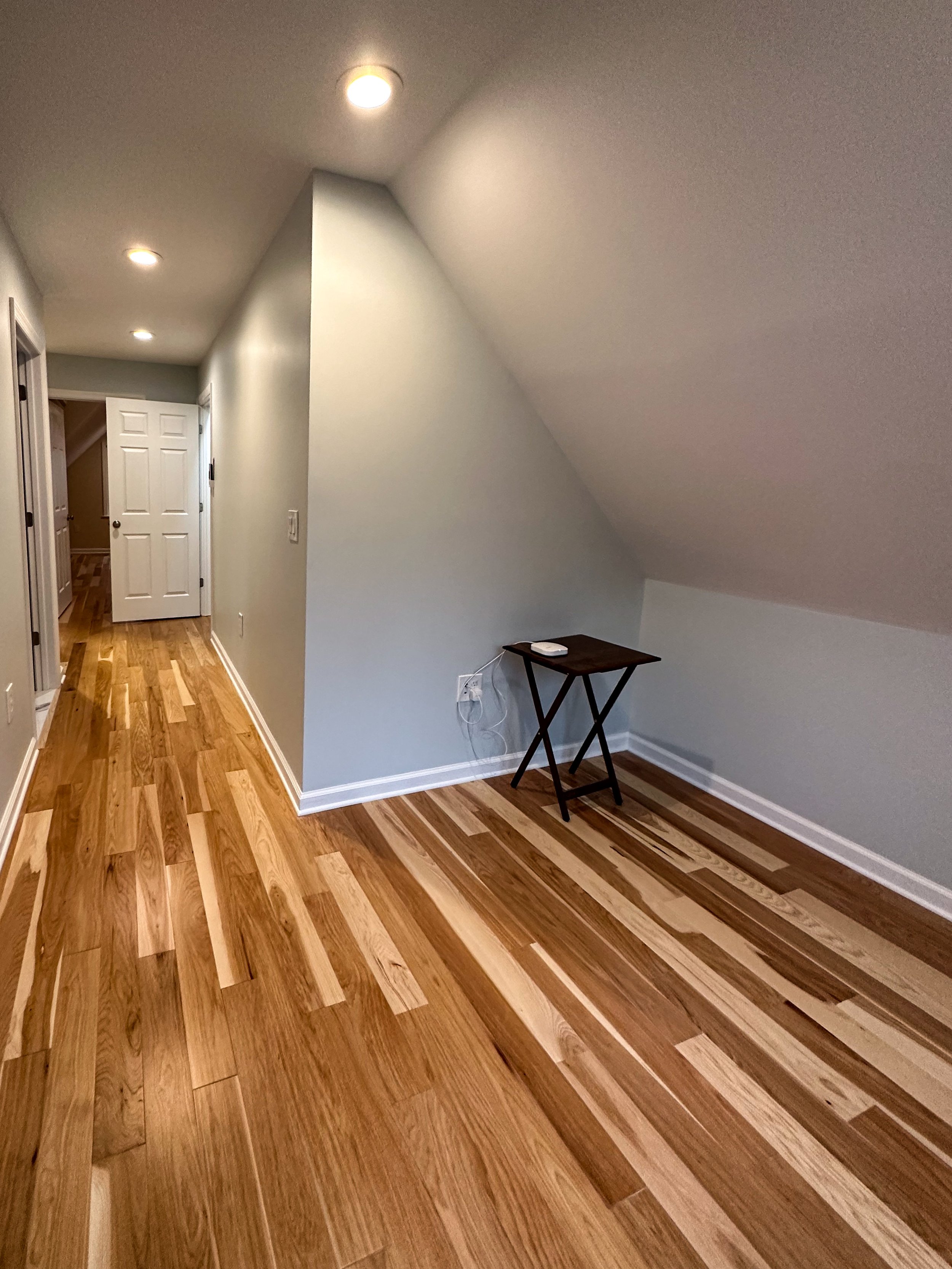 An enclosed room with sloped ceiling, hardwood flooring, plain white walls, some ceiling lights, an open door, and a small dark wood table with a device on top.