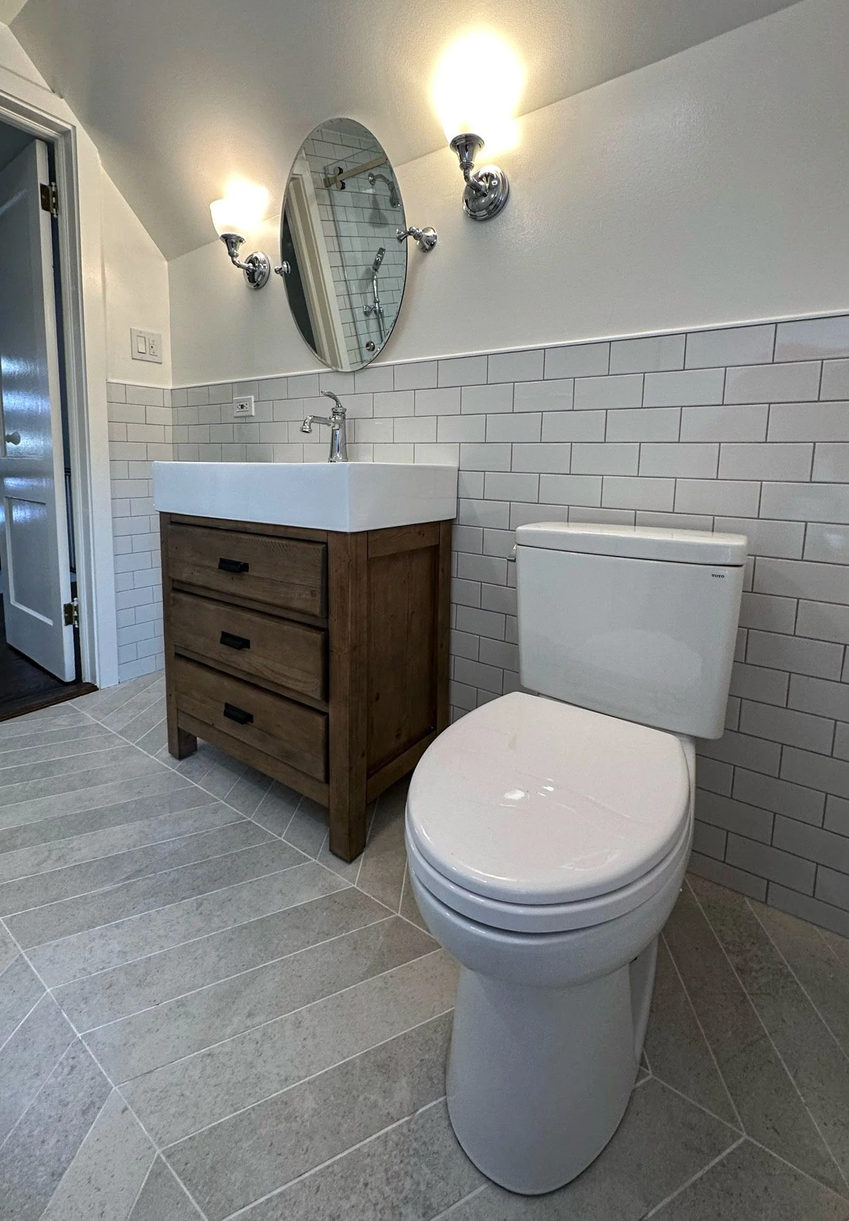A small bathroom featuring a white toilet, a rustic wooden vanity with a white sink, a round mirror, and wall-mounted light fixtures.