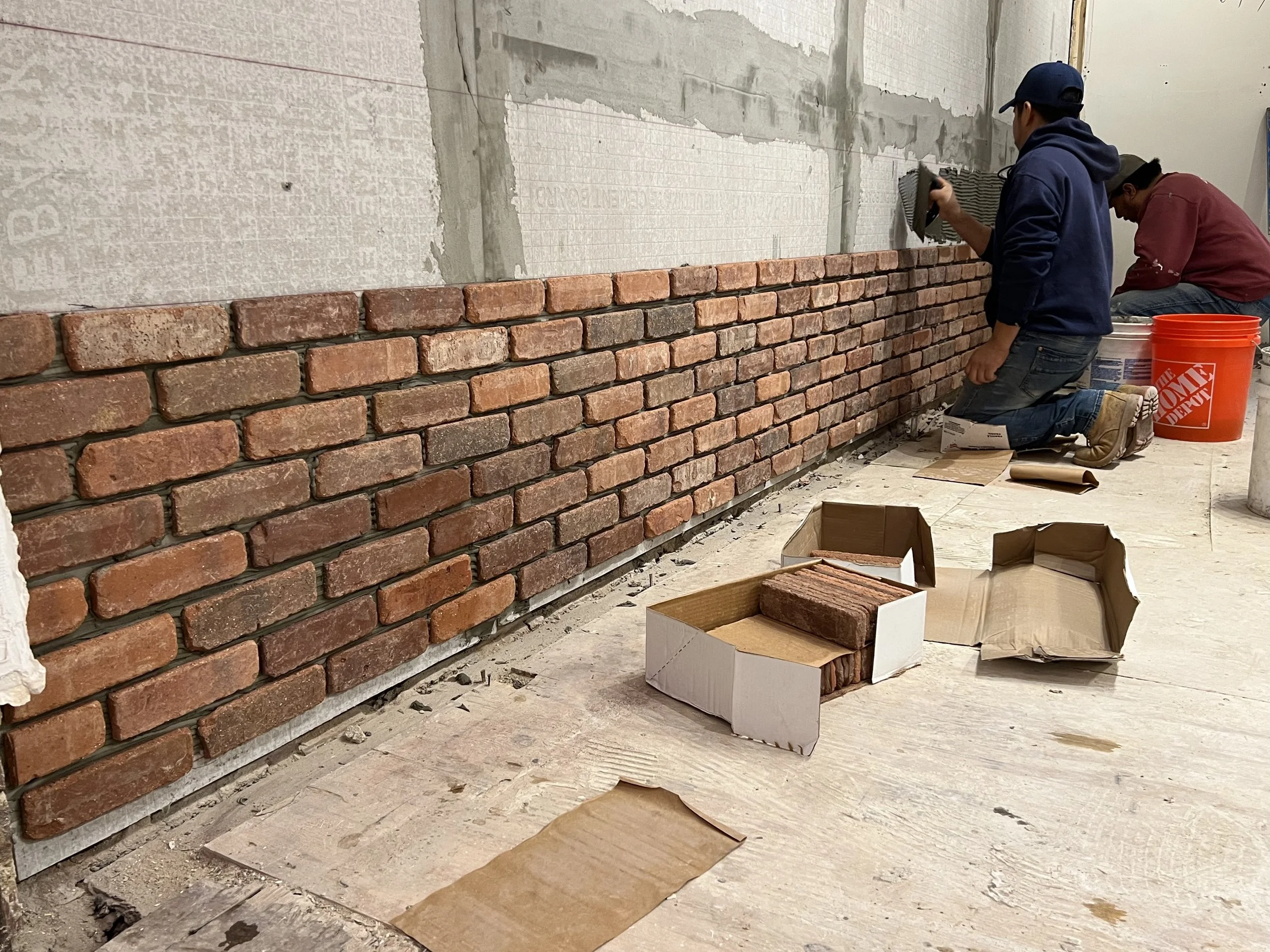 Two workers are installing a brick wall indoors. One kneels with a trowel applying mortar, while the other is also working on the wall. There are boxes of bricks and construction tools on the floor.