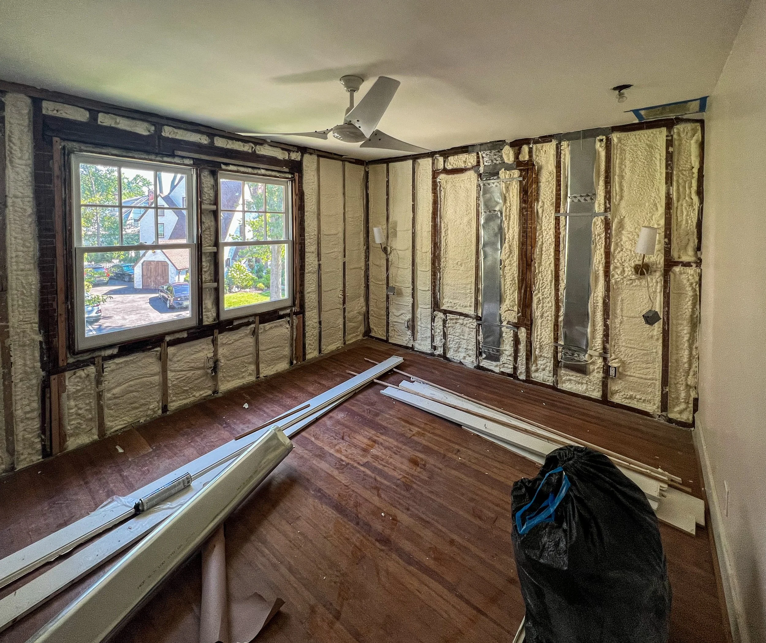 A room under renovation with exposed insulation on the walls, two new windows, and a ceiling fan. Construction materials and a black bag are on the wooden floor.