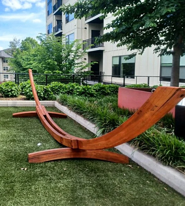Outdoor wooden lounge chair on a grassy area near plants and a pink planter, with a modern building and trees in the background.
