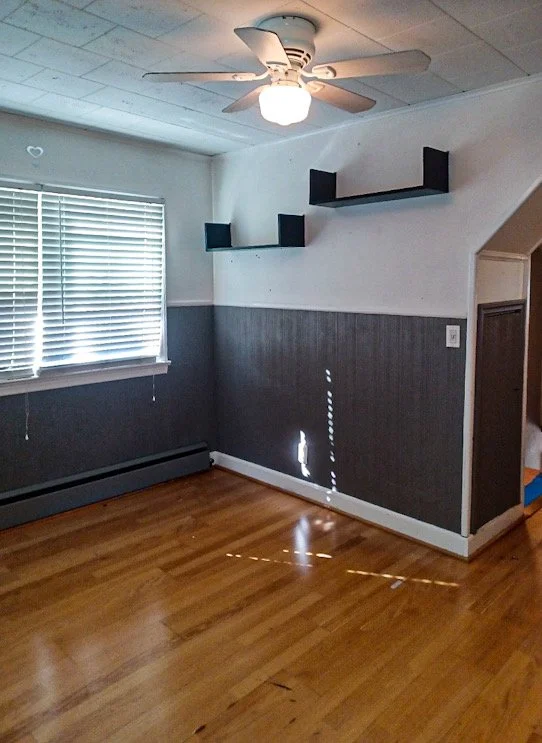 Empty living room with wood floor, gray wainscoting, window with blinds, ceiling fan with light, and black wall shelves.