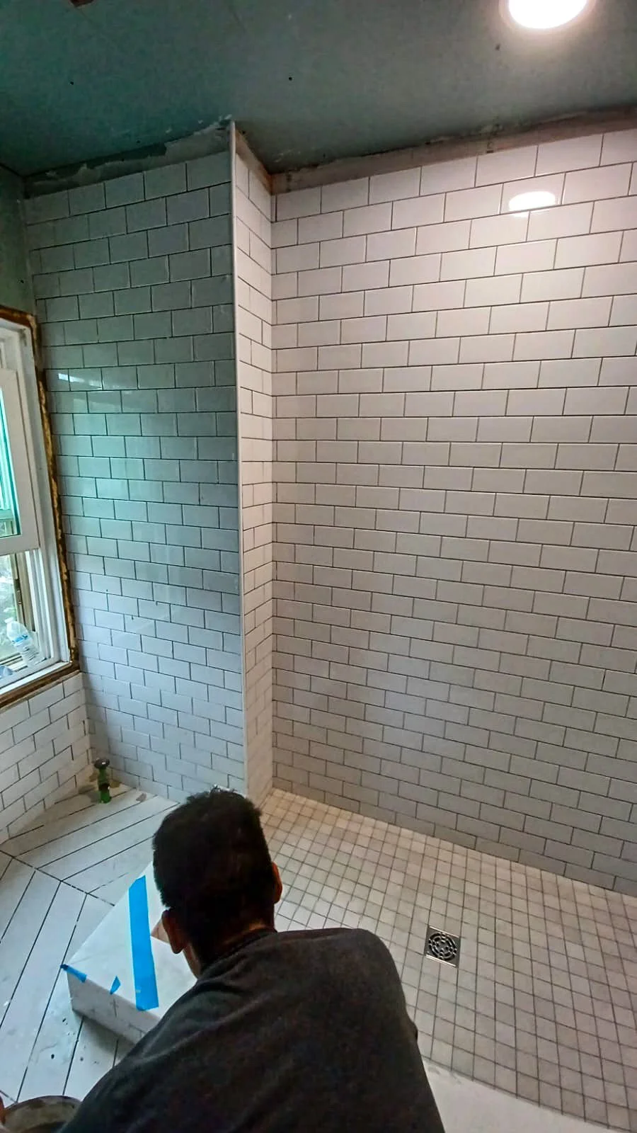 Interior of a bathroom with partially tiled walls and a tiled shower area. A person is working on the floor, and a window is visible on the left side.