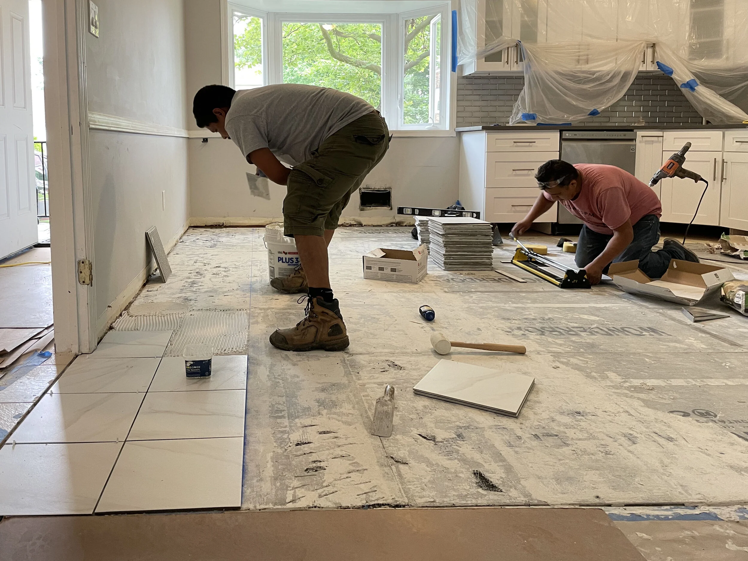 Two workers installing new white ceramic tiles on the floor of a kitchen under renovation, with tools and materials scattered around.