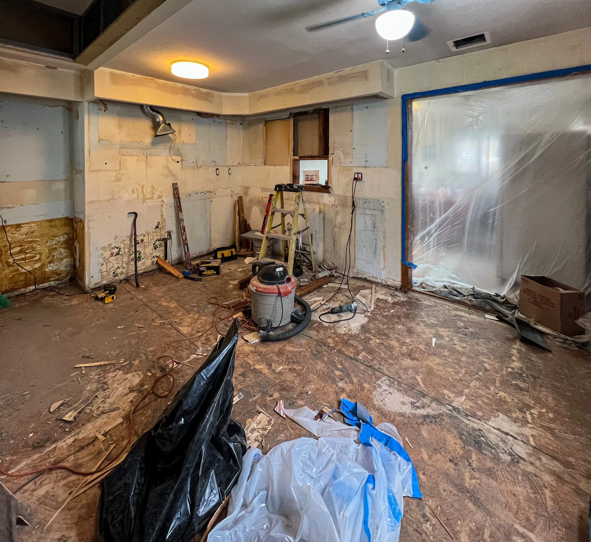 A room under renovation with exposed walls, construction tools, and debris on the floor, including a vacuum, ladder, and plastic sheeting.