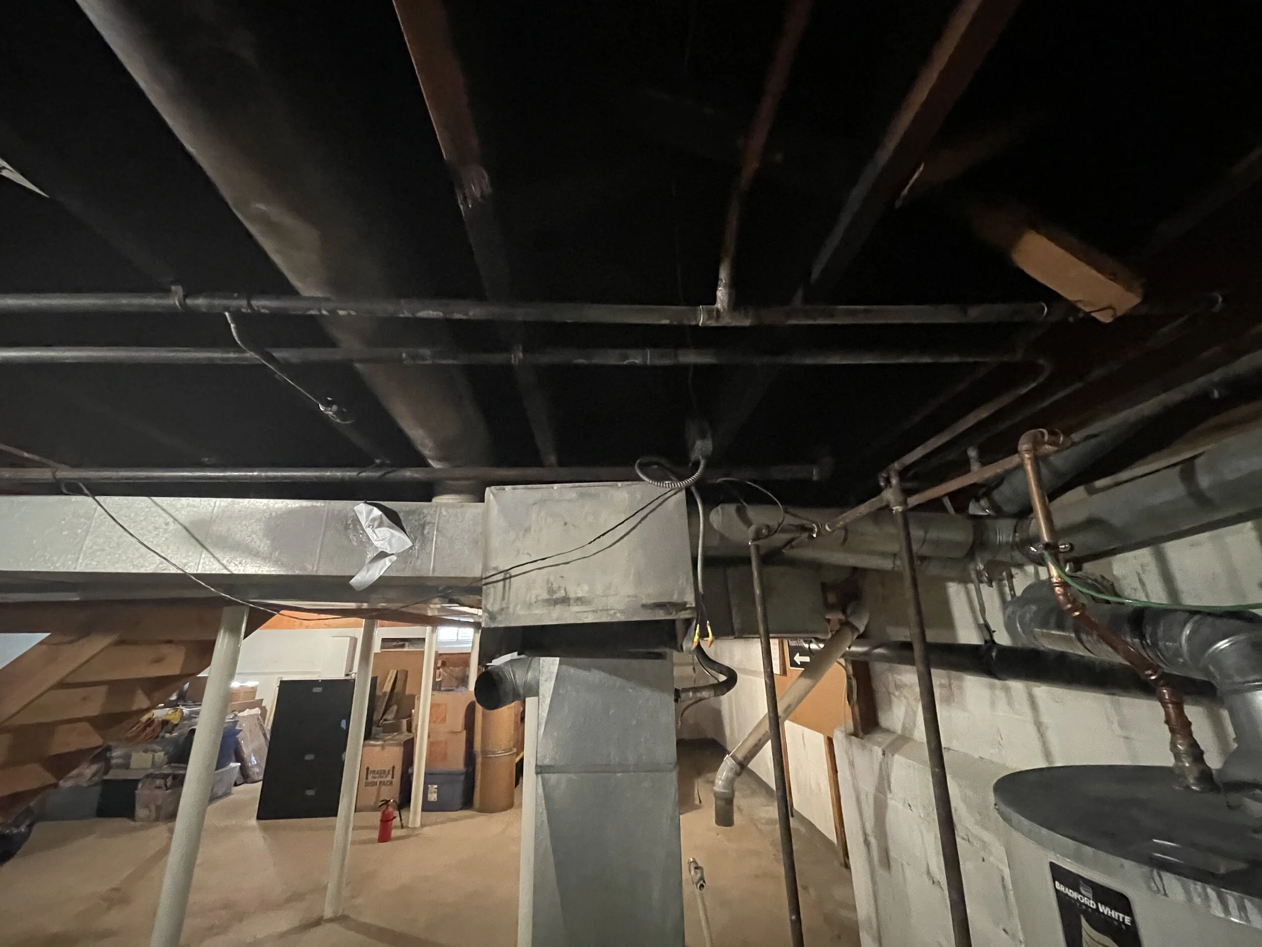 Basement with exposed pipes, ductwork, and wiring, unfinished concrete walls and floor, storage boxes and miscellaneous items in background.