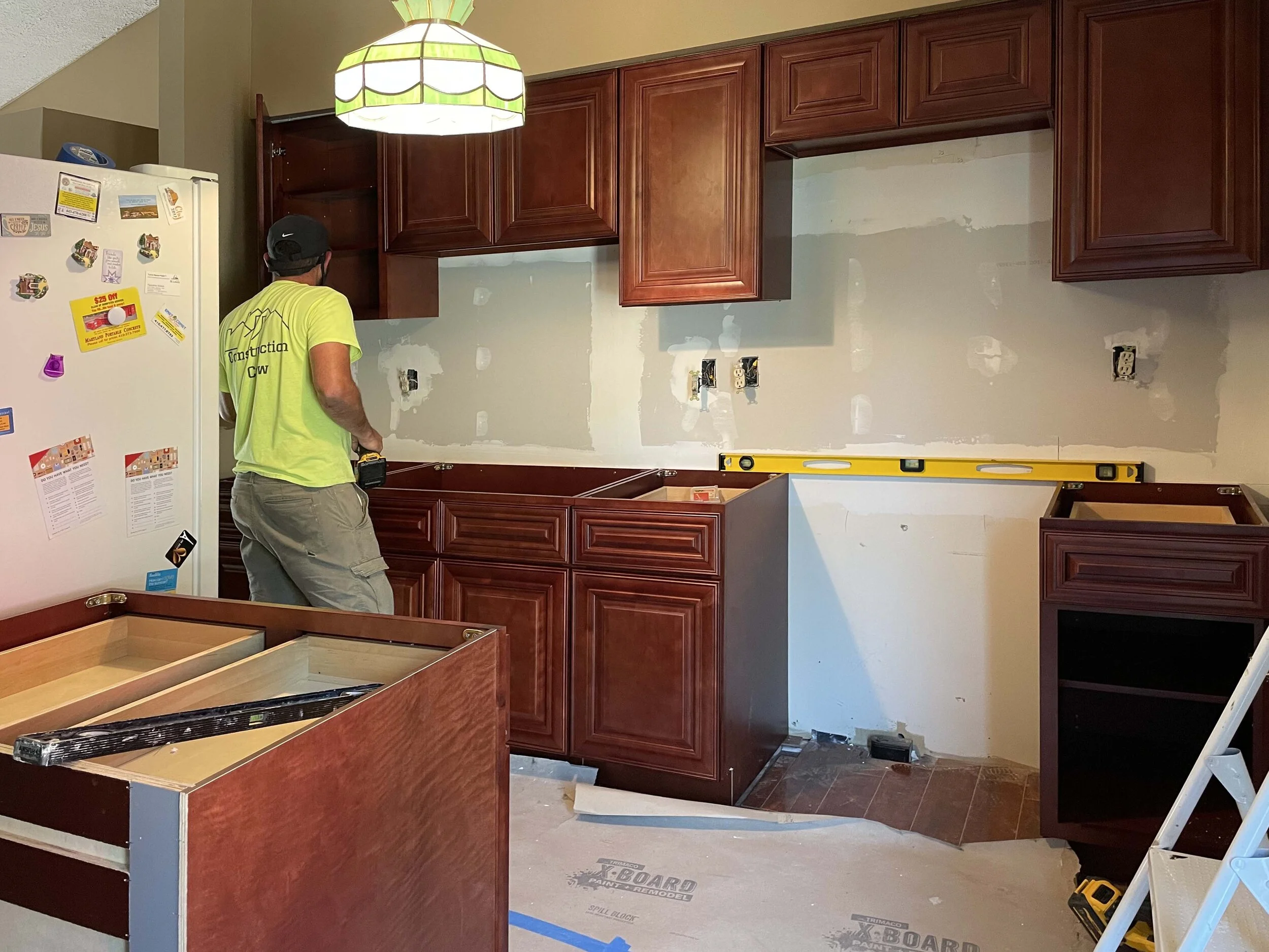 A man in a yellow construction shirt and gray pants working on kitchen cabinets during renovation, with partially installed cabinets, a level on a counter, and construction tools around.