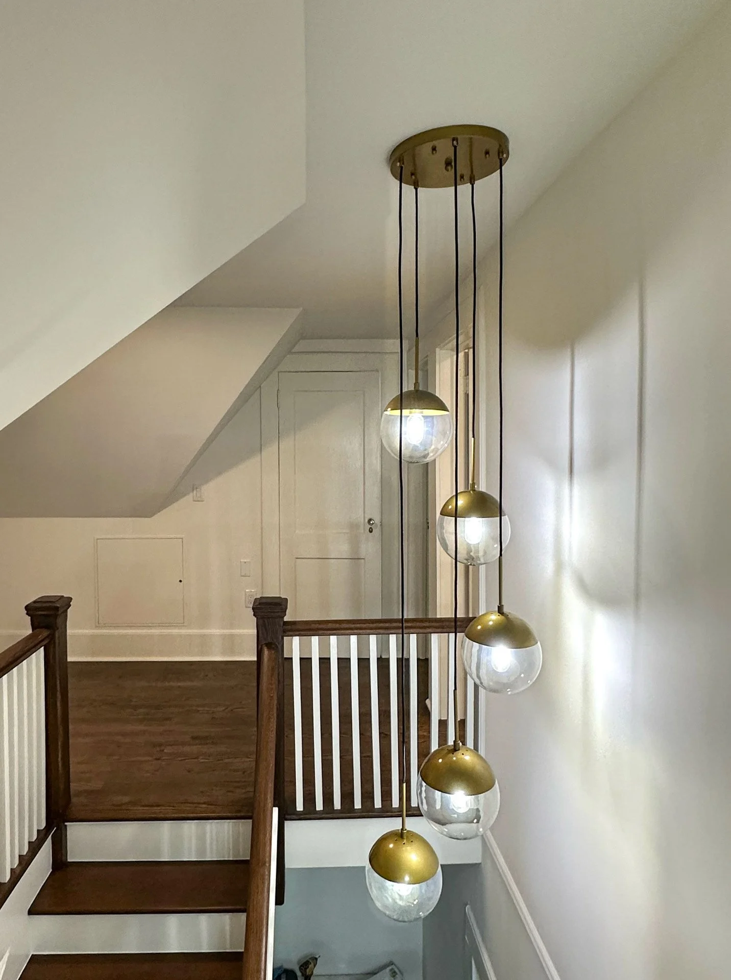 A modern hanging chandelier with five glass globes and gold accents hangs from a ceiling in a residential interior, illuminating a staircase with wooden handrails and white walls.