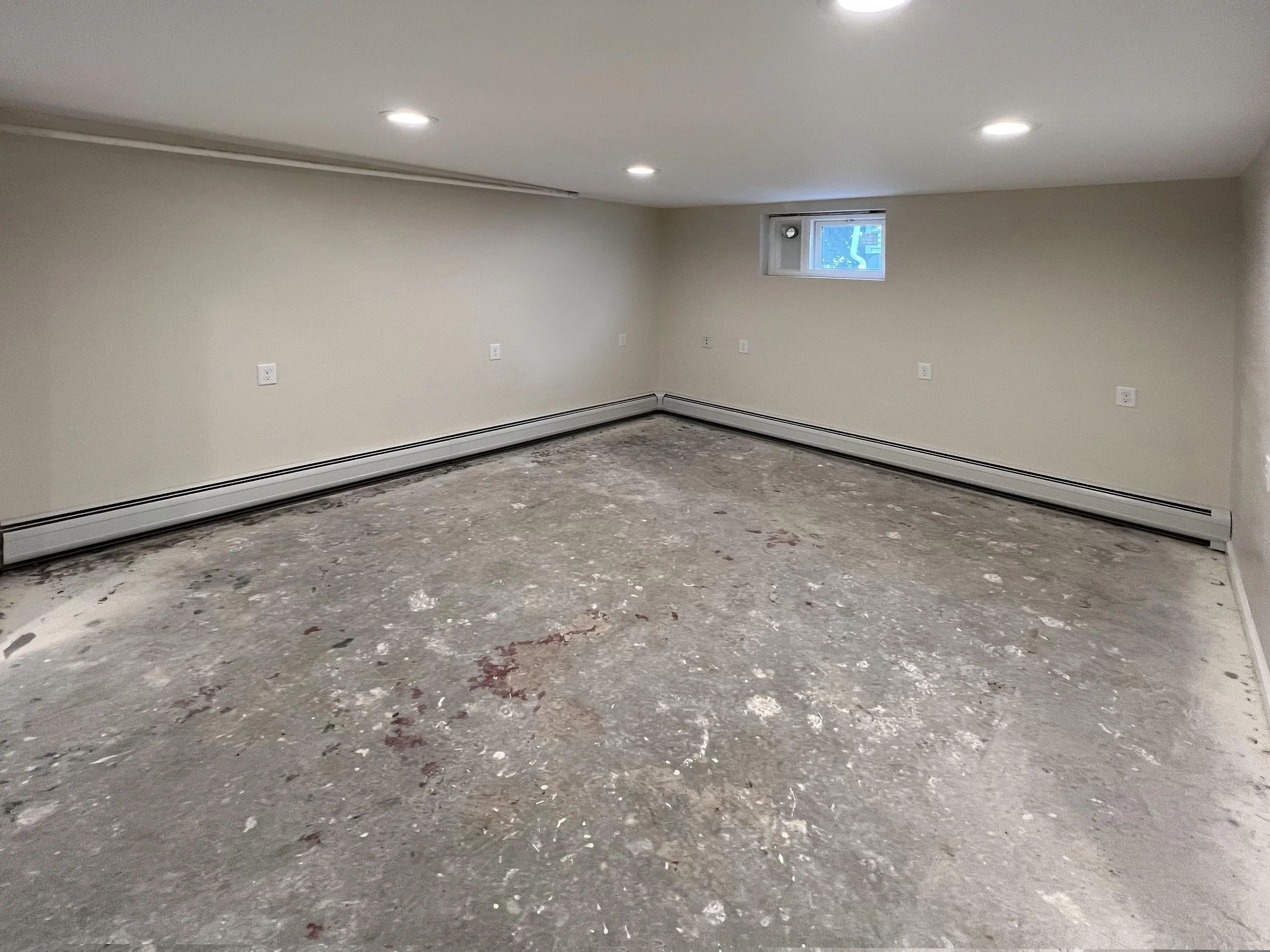 Undeveloped basement with concrete floor, beige walls, three recessed ceiling lights, a small window, and baseboard heater.