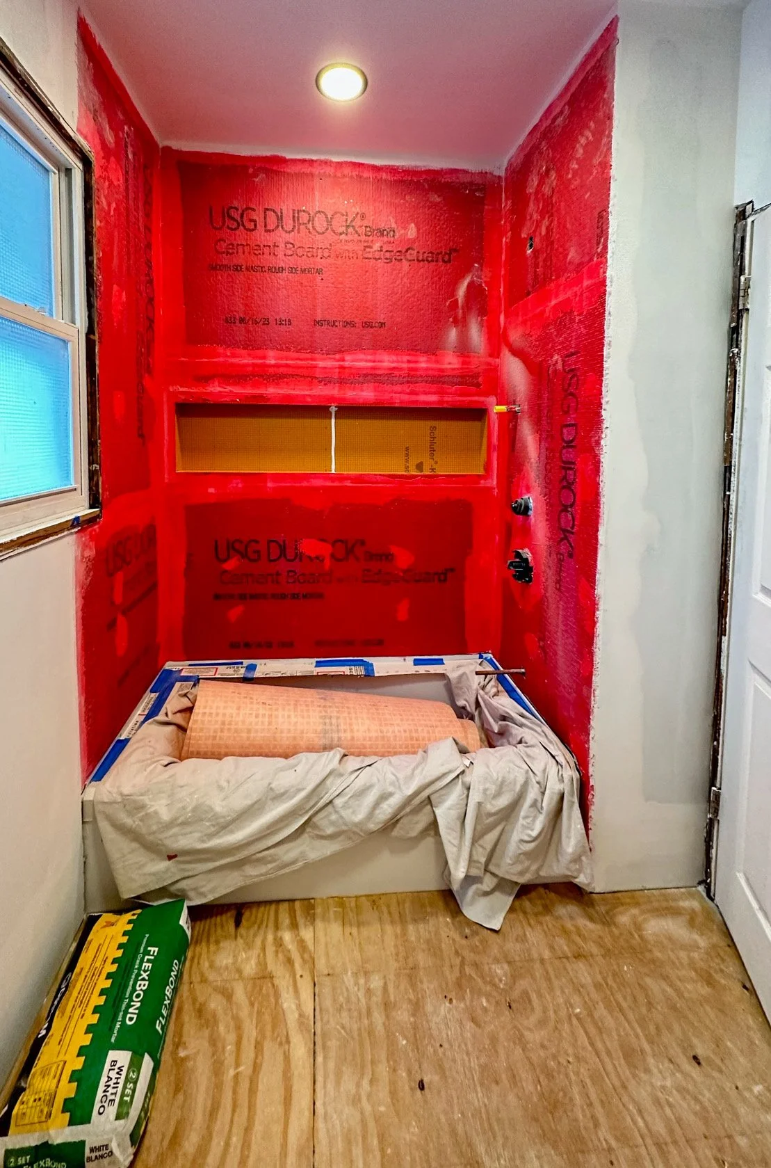 A small room under renovation with red cement board walls, a partially visible window, an unmade bed or cot with a pillow and sheets, a sheet of fabric or paper on top, a packed bag of insulation or building material on the wooden floor, and construc
