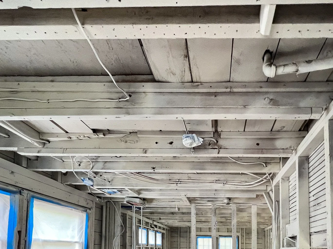 Interior view of a house under renovation with exposed ceiling beams, pipes, electrical wires, and construction materials, with partially covered windows.