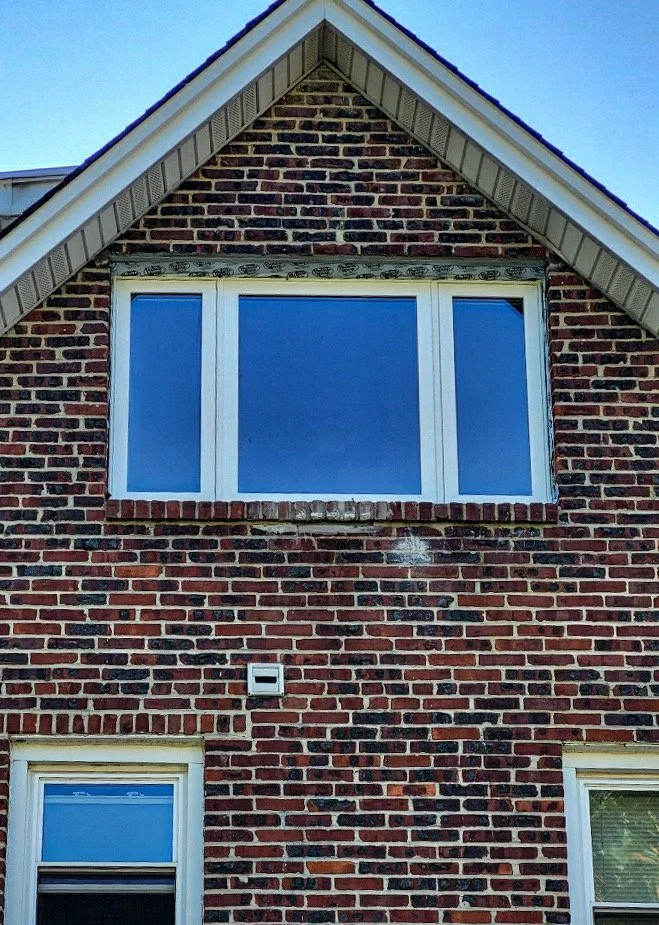 Close-up of the upper part of a brick house exterior, showing a gable roof with white trim, a large rectangular window, and brick wall details.