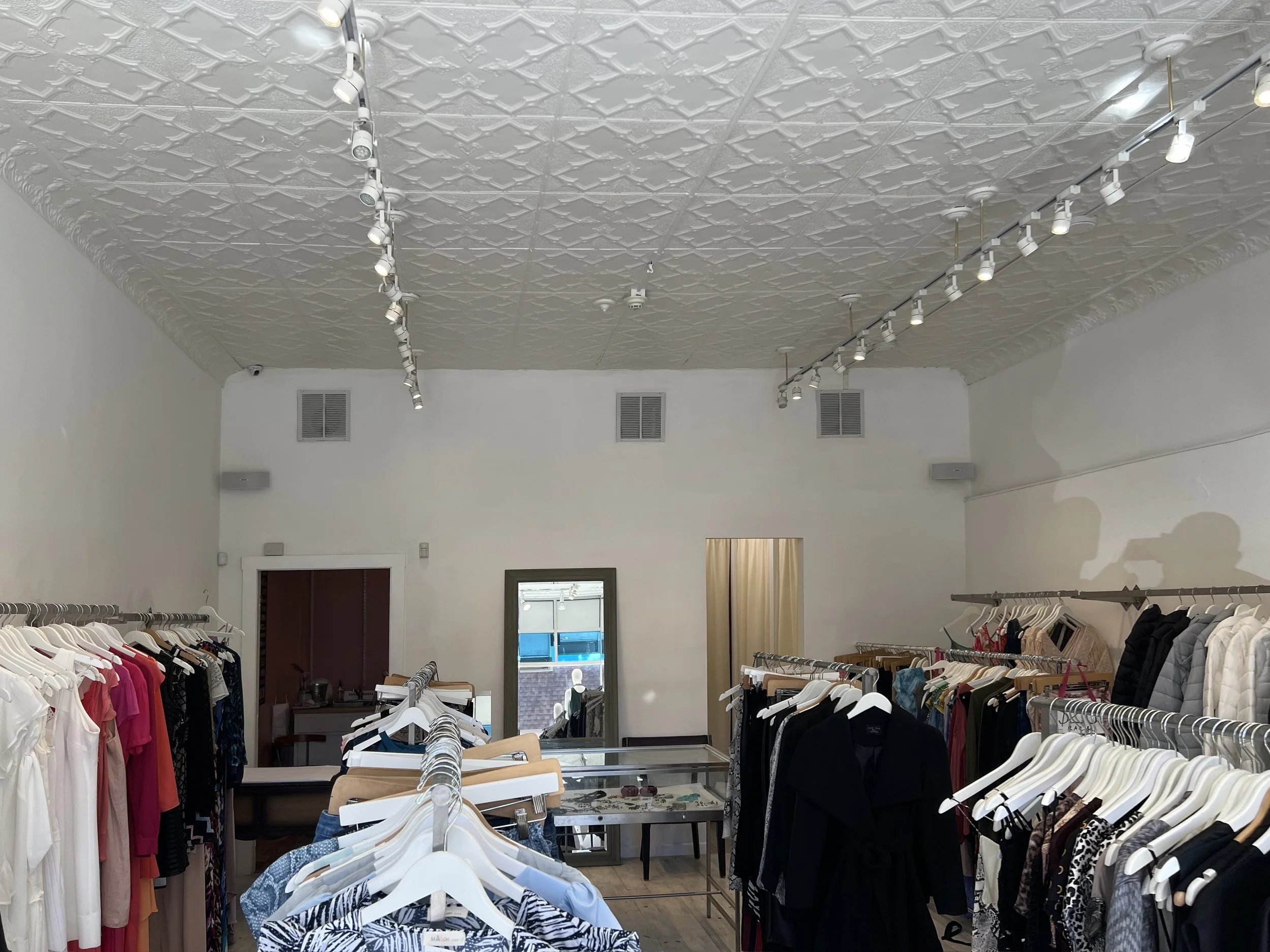 Interior of a clothing store with racks of dresses, shirts, and jackets, a large mirror, and ceiling lights.